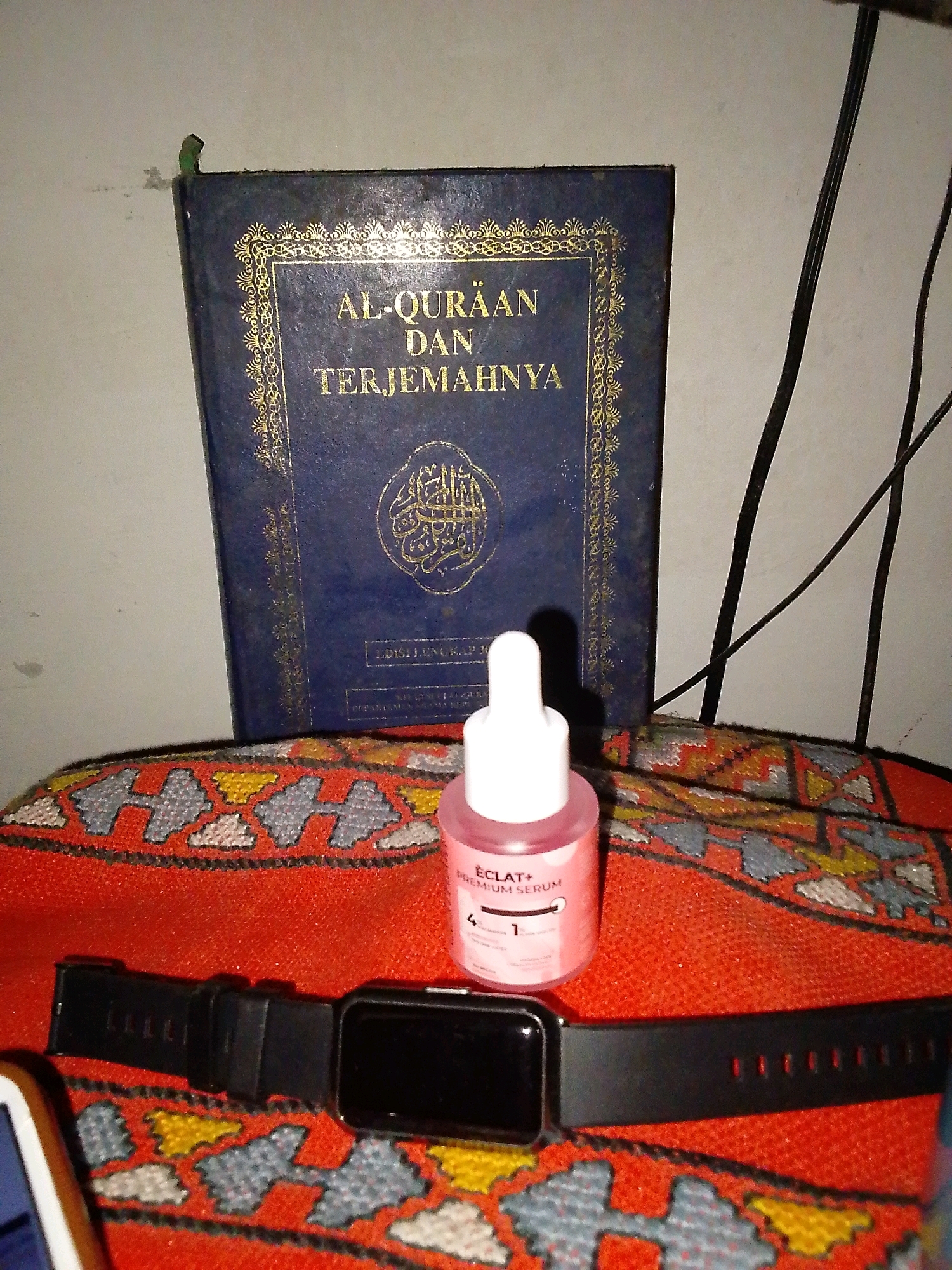 A close-up shot shows a book titled "Al-Qur'an dan Terjemahnya" in Indonesian, positioned vertically. To its right, a small bottle of "ÉCLAT+ PREMIUM SERUM" with a pink liquid and a white dropper cap stands on a red fabric with an orange and grey geometric pattern. Below the serum bottle and to the left, a black smartwatch with a digital display rests on the same patterned fabric. The background is a plain, off-white wall with two black cables running vertically. A small green tab is visible at the top left corner of the book's cover. The scene is indoors, with no specific city or location details identifiable from the image itself, other than it appears to be a personal space.
