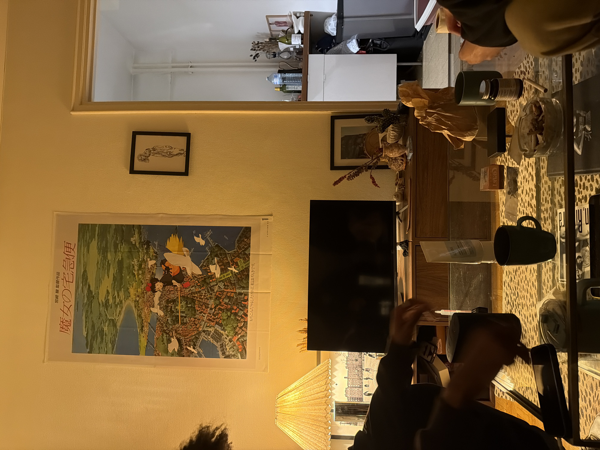 The image portrays an interior view of a living space located in Paris, France, characterized by warm, artificial lighting. A large, vertically oriented poster for "Kiki's Delivery Service" (魔女の宅急便) is prominently displayed on the left wall, depicting a witch flying over a detailed European-style coastal town. Below this, a black flat-screen television rests on a wooden media console. To the right of the poster, a smaller framed drawing of a slender, standing figure is mounted on the wall.
Two individuals are partially visible, seated around a glass coffee table. On the left, a person with dark, curly hair and wearing a dark long-sleeved garment holds a dark mug and gestures towards a smartphone on the table. On the right, another person's lower arm, hand, and a segment of their light-colored pants are visible as they manipulate a small object, appearing to prepare a cigarette.
The glass coffee table, situated over a leopard-print rug, holds several items: multiple dark mugs, a clear glass ashtray containing cigarette butts and ash, an unlit cigarette, a small glass jar (likely supplements), a clear plastic container with a lid, a bowl of snacks, a black portable speaker, a smartphone, a white tube of lotion, and a crumpled brown paper bag. The wooden media console displays decorative elements including vases, dried botanicals, and a framed picture.
Through an open doorway on the right, an adjacent room is visible, featuring white walls, exposed white piping, a white cabinet stocked with bottles (water, olive oil, wine), dried decorative items, and a dark trash can. The scene suggests a casual, domestic gathering.