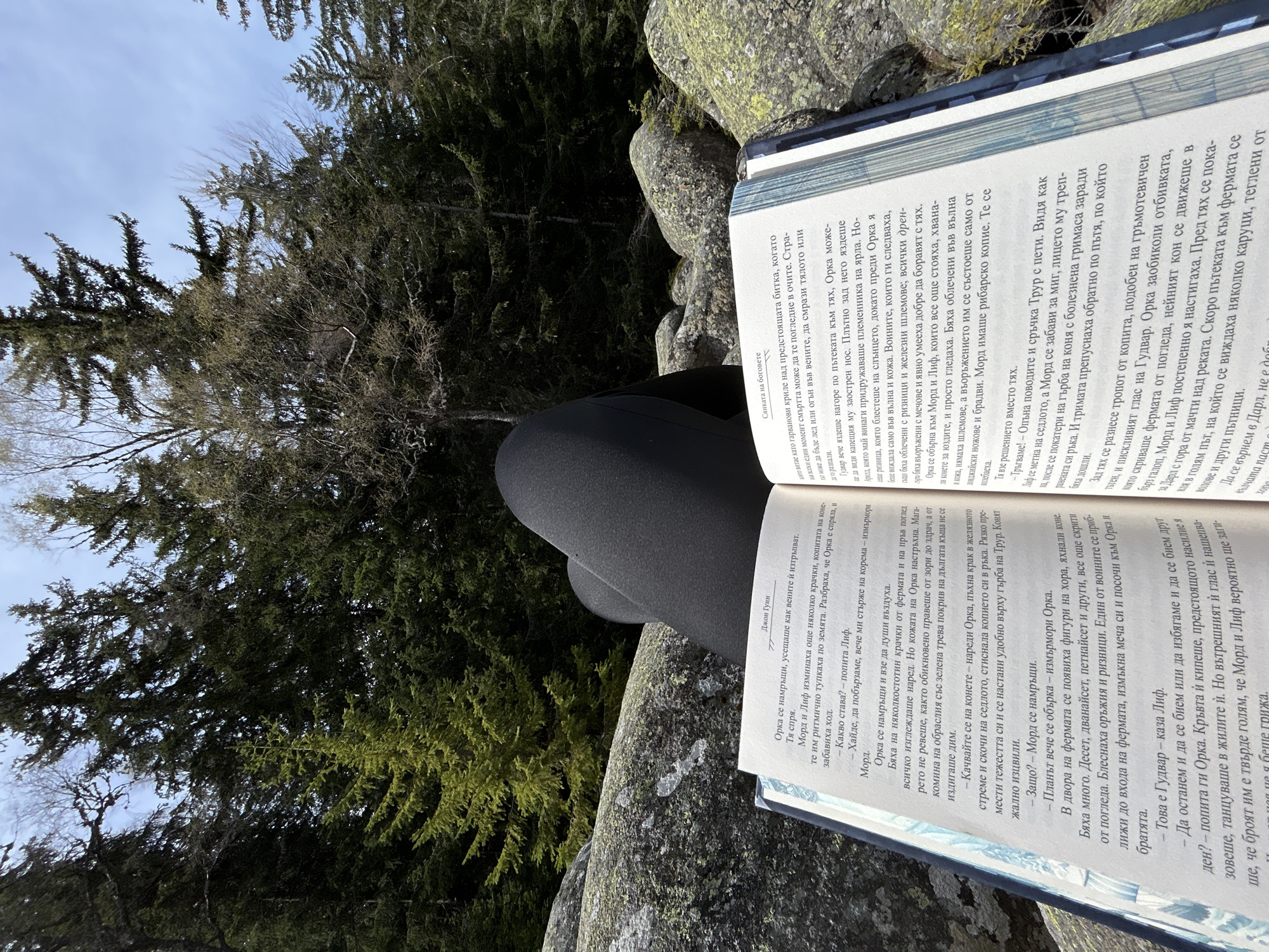 Here is a description of the image:
A person sits on a rocky outcropping, legs clad in black leggings, reading an open book. The book appears to be in Bulgarian. A forest of evergreen trees is in the background. The scene gives the impression of someone enjoying a quiet moment of reading in a natural setting.