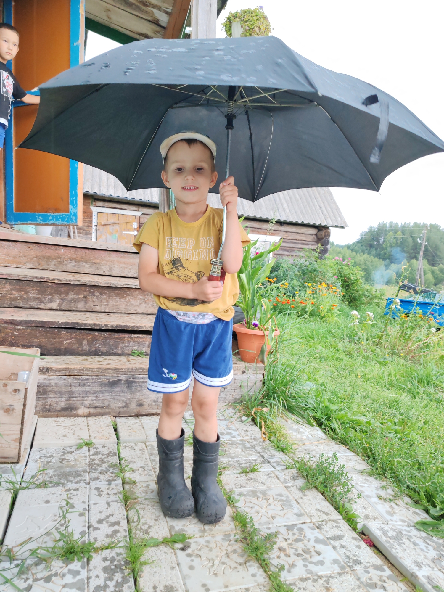 A young boy stands on a tiled porch in front of a wooden house in the village of Chuchlem, Russia, holding a black umbrella open. He is wearing a yellow t-shirt with a graphic and text that reads "KEEP ON DIGGING," blue shorts with white trim, and black rubber boots. The umbrella is tilted, with water droplets visible on its surface, suggesting it is raining or has recently rained. Behind the boy, wooden stairs lead up to a porch with an orange wall and blue trim. Further back, a log house with a corrugated roof is visible. The yard is overgrown with green grass and various flowers in pots. A second boy, wearing a black t-shirt, is partially visible on the porch to the left. In the background, trees and a utility pole are present. The overall impression is a rural, perhaps somewhat unkempt, setting during inclement weather.