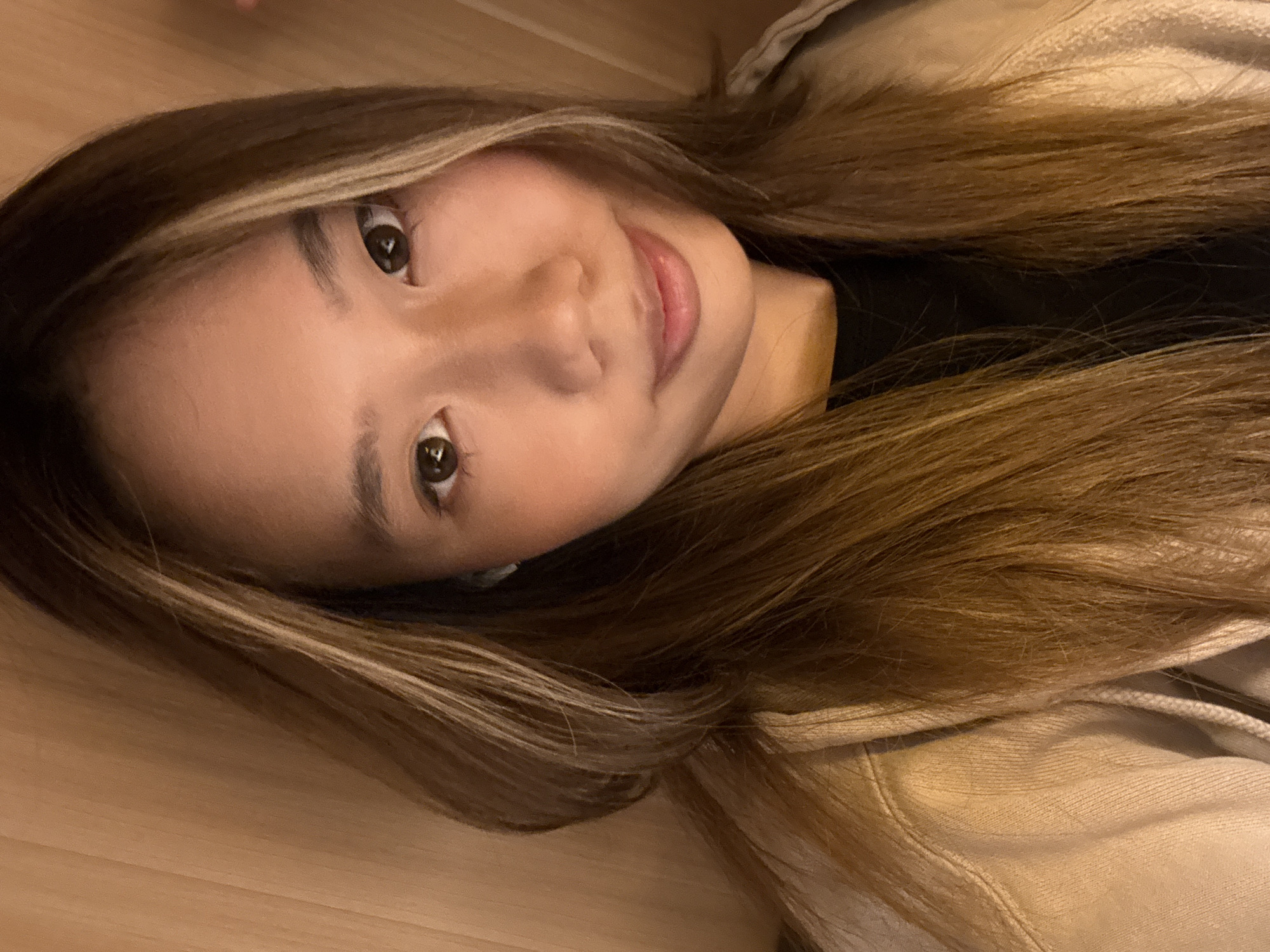 A young woman with brown hair with blonde highlights is smiling at the camera. She is wearing a black shirt and a tan jacket. The background appears to be a wood surface. There is no information available to determine if the image was taken in Yuzhong District, China.