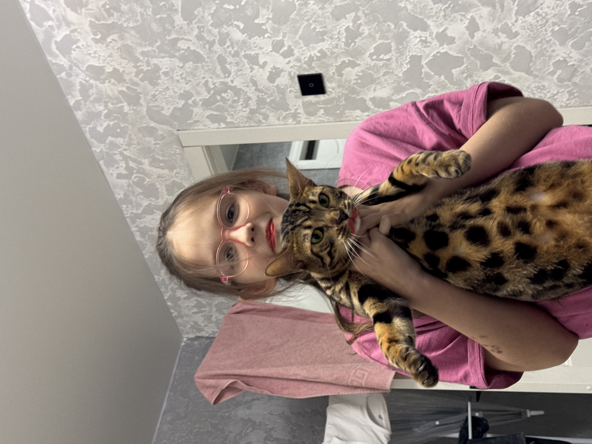 A young girl with glasses is holding a bengal cat indoors. The girl is wearing a pink shirt and has red lipstick on. The cat has a spotted coat. The background shows a textured wall and a doorway. A pink towel is hanging nearby. There are clothes on the floor. The city and region are not specified in the picture.