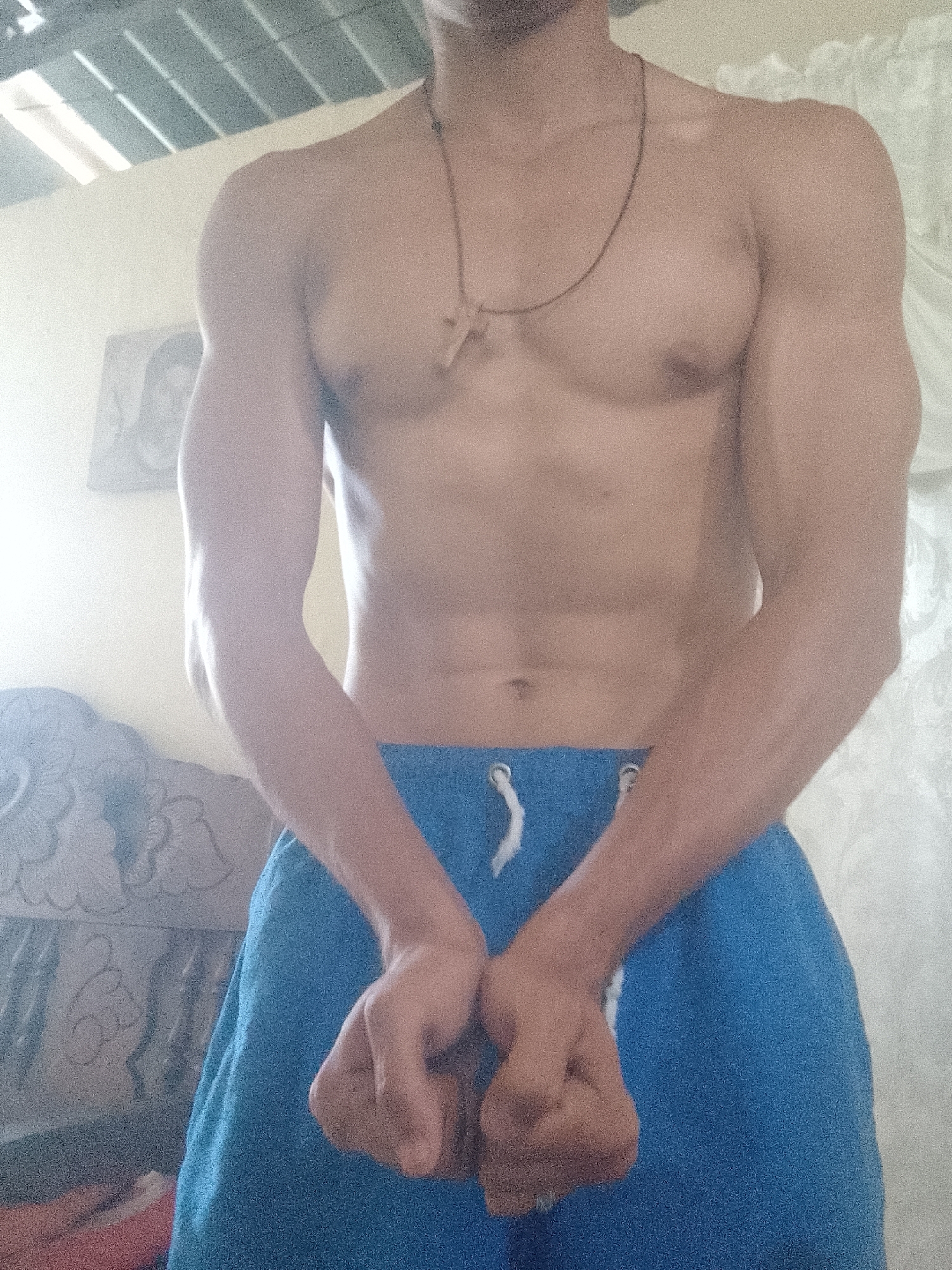 A shirtless, muscular man stands indoors, wearing blue shorts with white drawstrings. He has a cross necklace. His hands are clasped together in front of him. Behind him are a wall, part of a decorated wooden chair, and a window with sunlight coming through. The location is Juan Griego, Venezuela.
