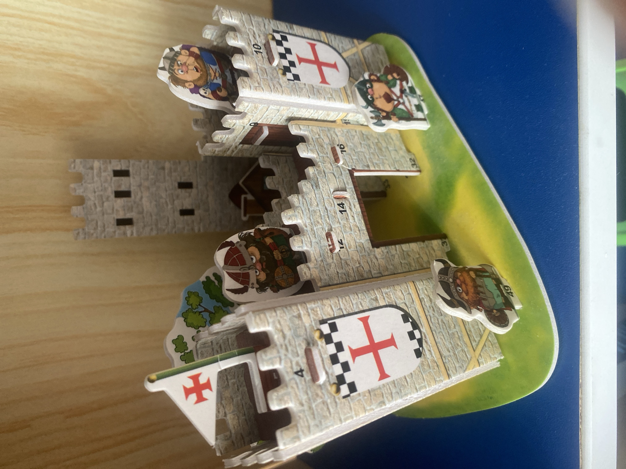 This is a close-up, eye-level, indoor shot of a 3D puzzle of a medieval castle. The puzzle is made of cardboard and features stone-like textures, battlements, towers, flags, and figures of knights and warriors. The castle is assembled on a green base that resembles grass, which is placed on a blue surface, possibly a table or shelf. In the background, a wooden surface with vertical lines is visible. The puzzle has several numbered sections, including 4, 10, 12, 14, 16, 32, and 49. The overall lighting is bright, suggesting it's daytime. There is no discernible location in the city of Vitsebsk, Belarus, within the image.
