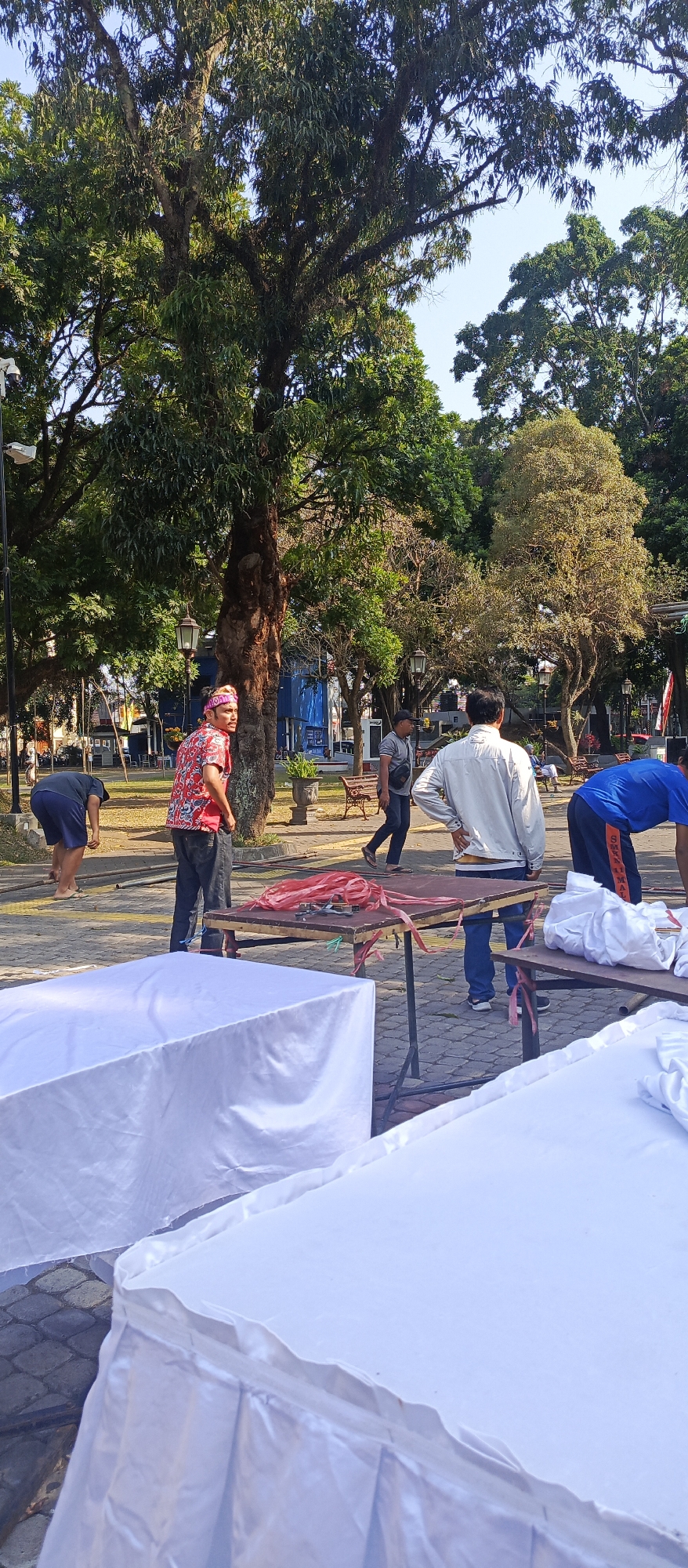 In a public park, likely in Malang, Indonesia, a group of people are engaged in an outdoor activity, possibly a community event or setup. In the foreground, two large tables are draped with white cloths, some appearing to be for food or merchandise. Red ropes are coiled on one of the tables. In the middle ground, several men are present. One man in a red and white patterned shirt and a headscarf stands near a tree, looking to the side. Another man in a light-colored jacket and blue jeans is walking away from the camera. A man in a blue shirt and dark pants is bending over near a table. In the background, more people are visible, along with park benches, trees, and some buildings. The overall setting suggests a daytime gathering in an open, green space.