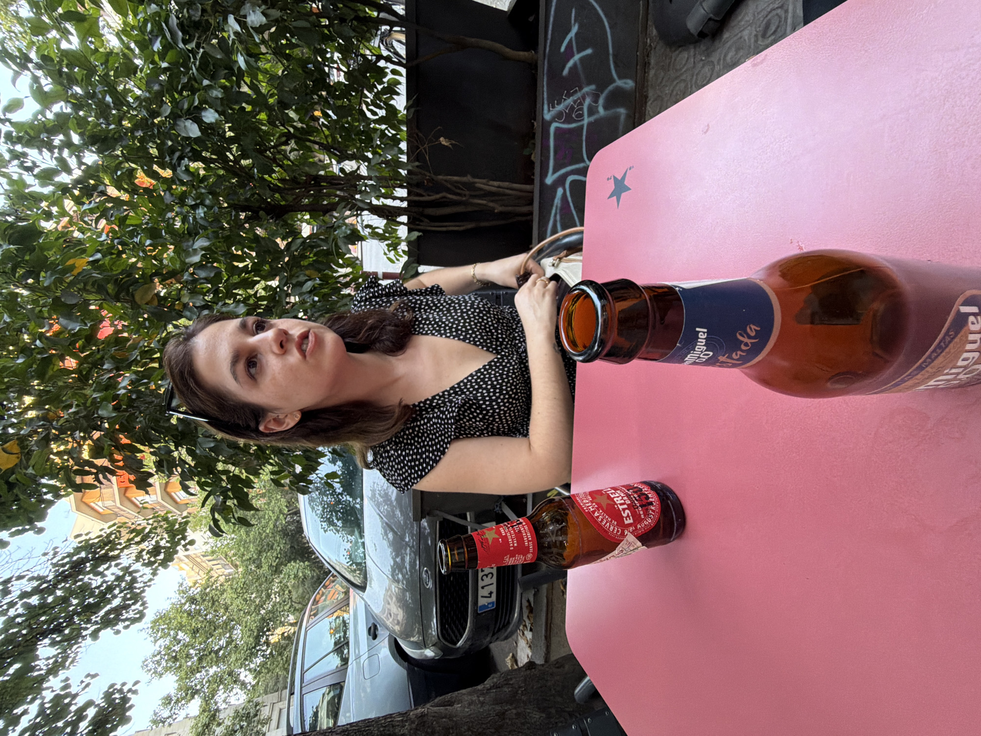 The image shows a young woman sitting at a pink table outdoors, possibly at a cafe or restaurant. She is wearing a black and white polka dot dress. Two beer bottles are on the table in front of her. One bottle, labeled "ESTRELLA DAMM" in red and blue, is mostly in the foreground. The other bottle, with a blue label that reads "CERVESA ME..." and "...DA DE MALTA, SENSE ARTIFICIALS...", is further back. A car is visible in the background behind the woman. Trees with green leaves are also present, suggesting an outdoor setting with some shade. The lighting appears to be natural, indicating it's daytime. The woman's expression is somewhat surprised or questioning. The scene feels casual and relaxed. The text "4137" is visible on the license plate of the car.