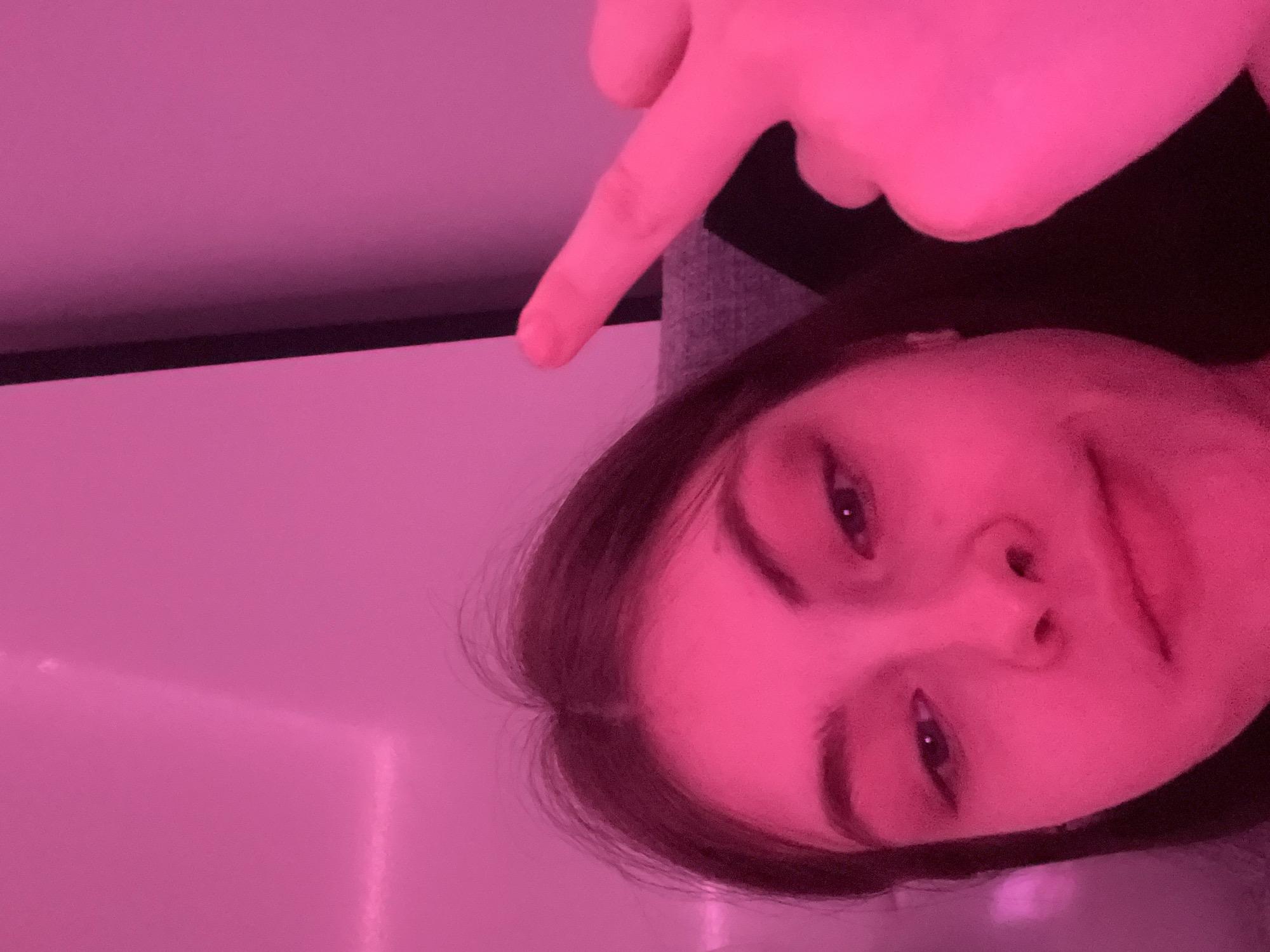 A close-up, low-angle shot of a person with fair skin, dark hair, and visible eyebrows, eyes, nose, and lips, is illuminated in a pinkish light. The person's right hand is visible at the top, with the index finger pointing downwards towards the person's face. The background appears to be a wall, also illuminated in pinkish light. The location is not discernible.