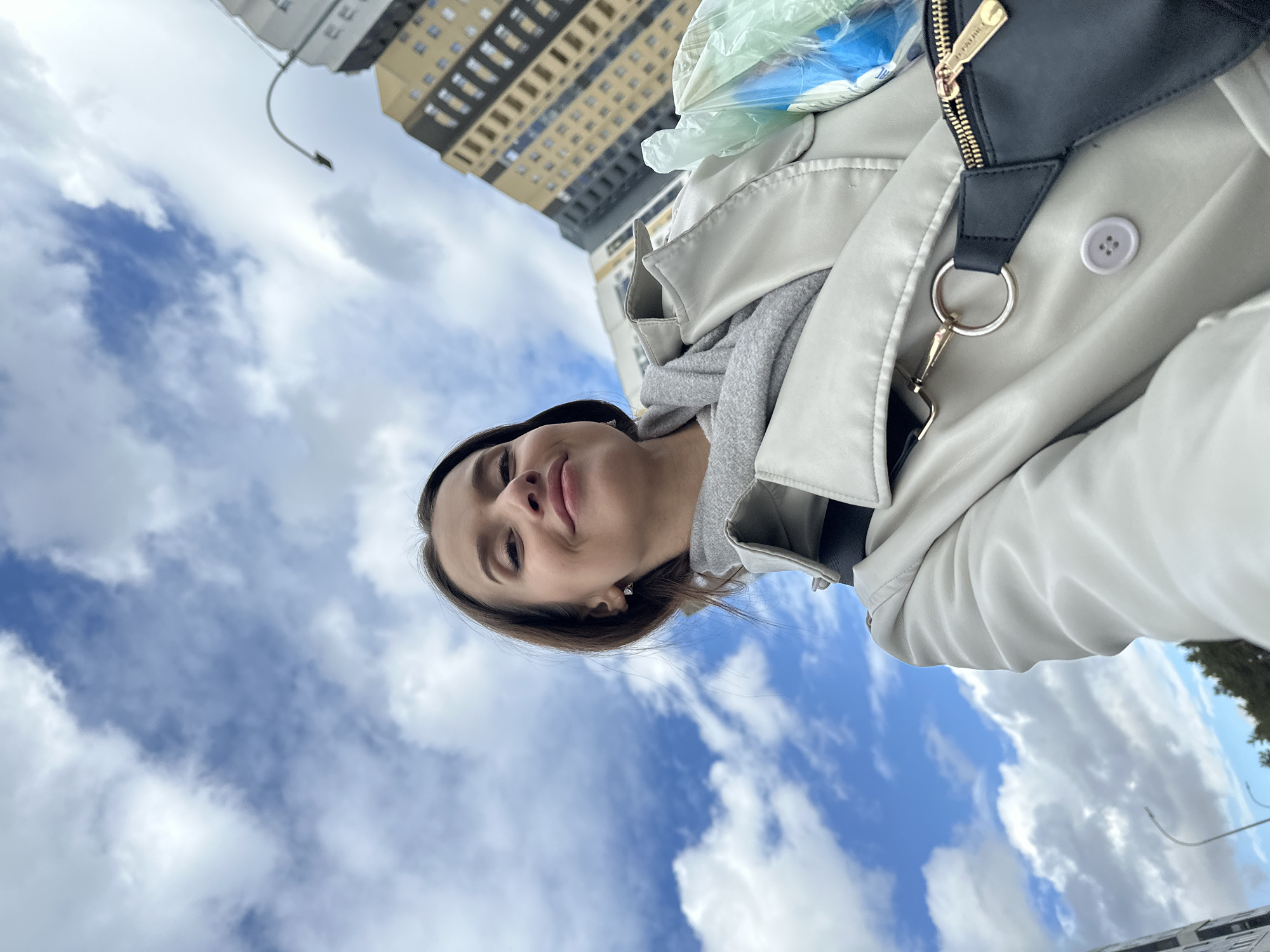 A low-angle shot captures a woman wearing a light-colored jacket and a grey hooded sweatshirt. A black purse with a gold zipper is visible on her shoulder. Above and behind her is a cloudy blue sky and the upper portion of a tall apartment building. This image was likely taken in Yekaterinburg, Russia.
