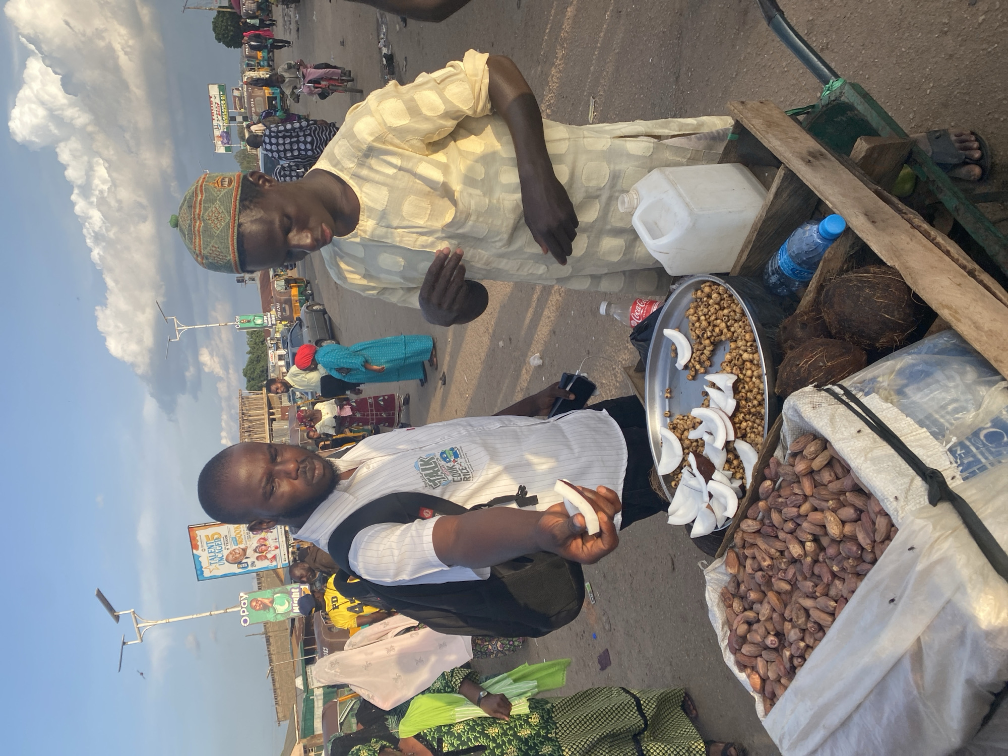 This image captures an active street market scene in Gura Top, Nigeria, under a partly cloudy sky. In the foreground, two men are engaged in a transaction at a mobile vendor's cart.
The man on the left, a customer, has dark skin, a short beard, and wears a white striped collared shirt with a black backpack. His right hand is extended, holding a crescent-shaped piece of white coconut, seemingly offering it or showing it to the vendor. His gaze is directed downwards. A logo reading "TALK COOK RICE" with cartoon characters is visible on his shirt.
The man on the right, the vendor, also has dark skin and wears a green and beige patterned kufi cap. He is dressed in a light yellow tunic with a raised, patterned texture. His hands are positioned over the cart, and his head is slightly bowed, looking towards the items on display.
The vendor's cart, constructed from wood with a green metal frame and handle, holds various goods. A large white sack in the foreground is filled with numerous brown dates. On a silver metal tray, there are many small, round, light brown nuts (possibly tiger nuts) mixed with several pieces of white coconut. Below the tray, within the wooden cart structure, two whole brown coconuts are visible, alongside a blue-capped plastic water bottle, a red-capped Coca-Cola bottle, and a white plastic jerrycan. The cart rests on an unpaved, brownish-red ground surface.
In the mid-ground and background, numerous people are visible walking and standing, some wearing traditional attire. Several three-wheeled auto-rickshaws (keke napep) and cars are present on what appears to be a busy street. A child is riding a pink bicycle in the distance. Buildings of varying heights, some under construction with visible scaffolding, line the street. A tall pole with solar panels for lighting stands prominently against the blue sky, which features scattered white cumulus clouds. Multiple advertising billboards are visible, including one green OPay sign and a blue banner advertising "TALENT UNCAGE D 5 YEARS" with an "$500K" prize.