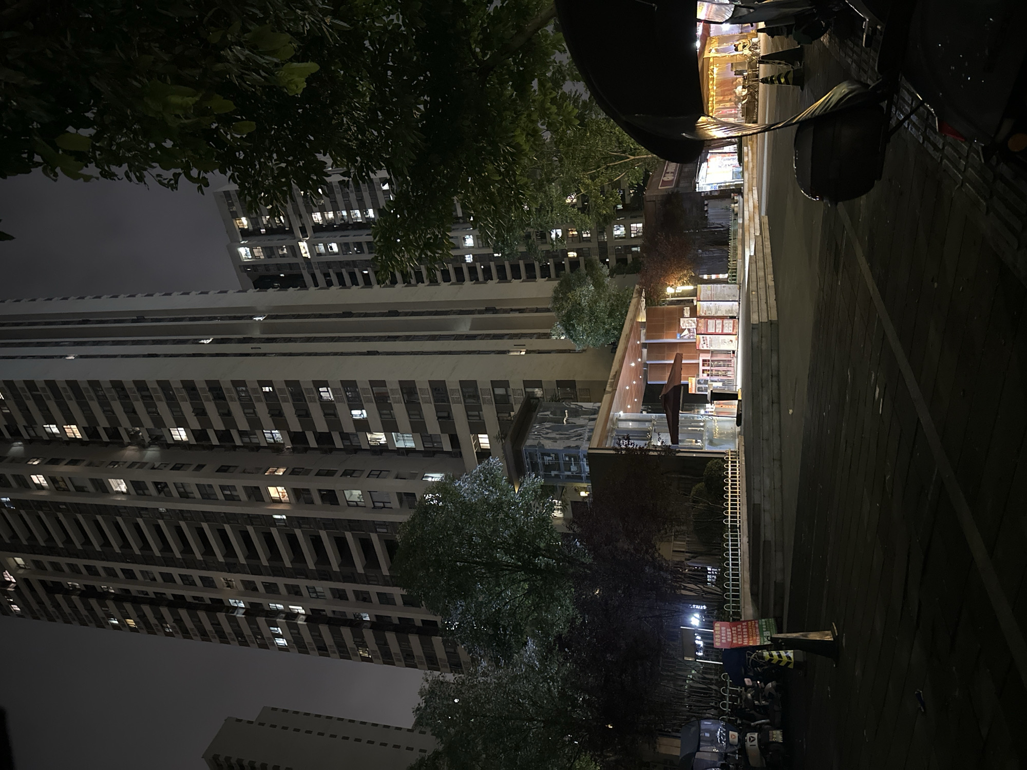 An outdoor night scene is depicted in Chongqing High-tech Zone, China. A large, multi-story residential building dominates the background, with lights visible in many of its windows. In front of the building, there are trees and a low-rise structure with an illuminated entrance and signage. Several parked electric scooters are visible on the left side of the frame, near a signpost. The foreground shows a paved area with dark wooden planks. On the right side of the foreground, a portion of a vehicle, possibly a three-wheeled electric scooter, is visible with a black canopy and a side mirror. Orange traffic cones are placed in the scene. The overall atmosphere is dark and urban.