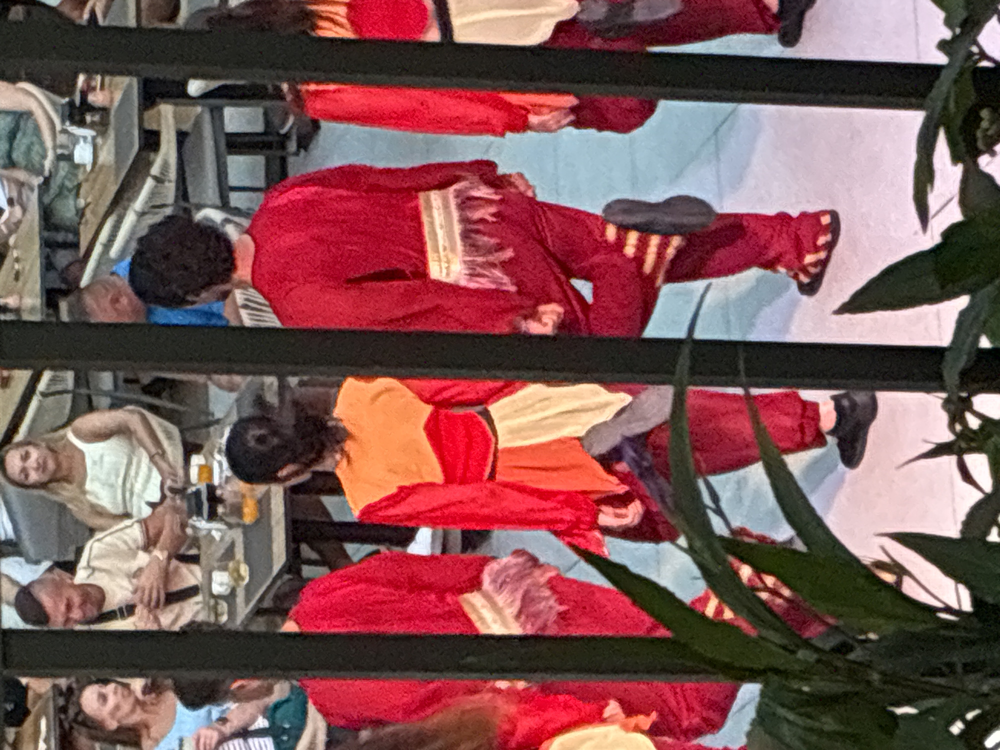 The image depicts an entertainment scene in Alanya, Turkey, viewed through dark vertical bars and partially obscured by green foliage in the immediate foreground. In the midground, five individuals, presumed to be performers, are visible from behind, dressed in elaborate costumes. These costumes are predominantly red, with one incorporating an orange upper garment and layered fabric around the waist. Specifically, the third performer from the left wears an orange V-neck top with red sleeves and a multi-layered, light-colored lower garment, with their right foot elevated in a pose. The fourth performer from the left is clad in a red costume featuring a golden-yellow horizontal waist belt with light-colored fringes, and their right leg is lifted and bent, displaying striped footwear.
In the background, multiple patrons are seated at tables within a venue resembling a restaurant or bar. Notable audience members include a woman with brown hair in a light blue top and a man with a distinctive haircut in a light-colored shirt, both seated at a table on the left. Further back, another woman with blonde hair in a white tank top is visible. Tables hold beverages, including one glass with an orange liquid. The background flooring is light in color. The performers' postures suggest active movement, indicative of a dance or theatrical performance.