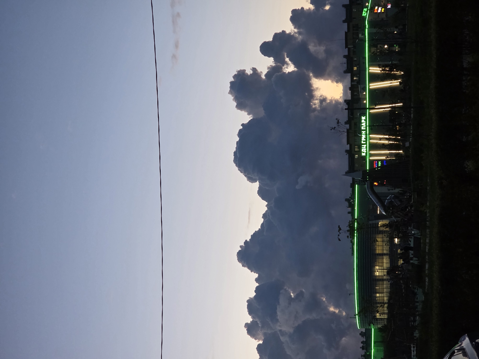 The image depicts a modern building with green neon lighting, identified by the signage "КДЦ ГРИН ПАРК" (KDC GREEN PARK), located in Shushary, Russia. The building appears to be a cultural or entertainment center. Behind the building, dramatic, dark cumulonimbus clouds are illuminated by a setting sun, creating a striking contrast against the twilight sky. A thin power line stretches horizontally across the upper portion of the frame. The foreground shows a dimly lit area with some trees and what appears to be a park or outdoor space, with a glimpse of a vehicle on the far left. The overall scene suggests a transition from day to night, with the artificial lights of the building standing out against the natural, yet imposing, cloud formations.