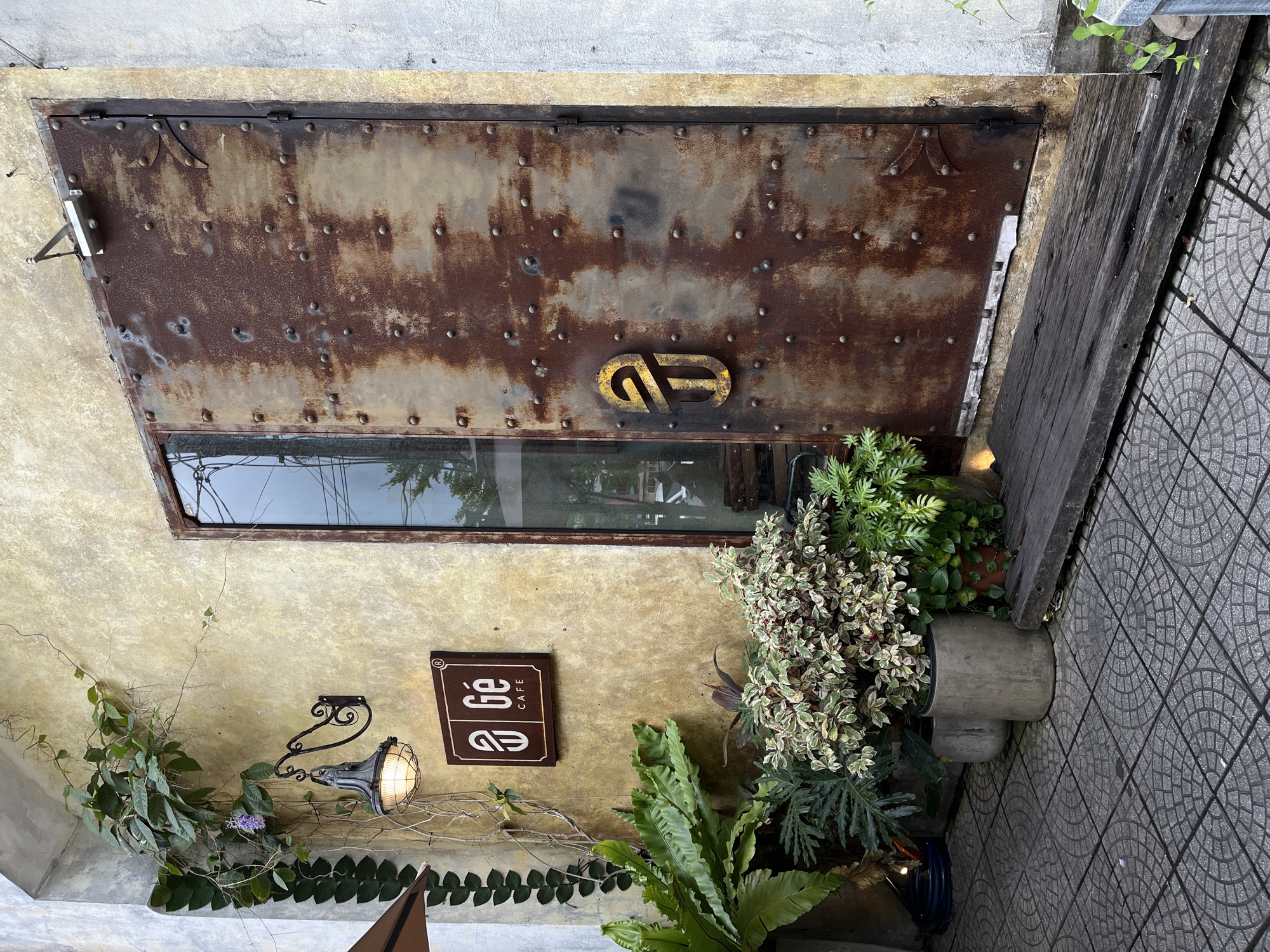 The image captures a rustic and charming entrance, likely a cafe or shop, situated in Đà Nẵng, Vietnam. The scene is dominated by a heavily rusted metal door, studded with numerous small rivets, suggesting an aged or industrial aesthetic. A rectangular glass panel is integrated into the lower half of the door, reflecting the outdoor environment. Prominently featured on the upper section of the door is a golden, stylized symbol, possibly an intertwined "G" and "E" or "C", matching the cafe's branding.
To the left of the door, a textured, light yellowish-beige wall is adorned with lush green foliage, including climbing vines and various potted plants, such as large ferns and plants with variegated leaves. A vintage-style black metal wall sconce with a caged bulb emits a warm, inviting glow, casting light on the wall and plants. Below this light fixture, a dark brown rectangular sign with white lettering clearly displays the text "GÉ CAFE" in a stylized font.
The ground to the right of the entrance is paved with grey tiles featuring a distinctive semicircular pattern. A weathered wooden beam or plank leans against the doorframe on this side, complementing the rustic theme. The overall ambiance is one of natural warmth and aged character, typical of an artistic or cozy cafe setting. No people are visible, and the time of day appears to be evening or dusk, given the illuminated sconce, though natural light is still present.