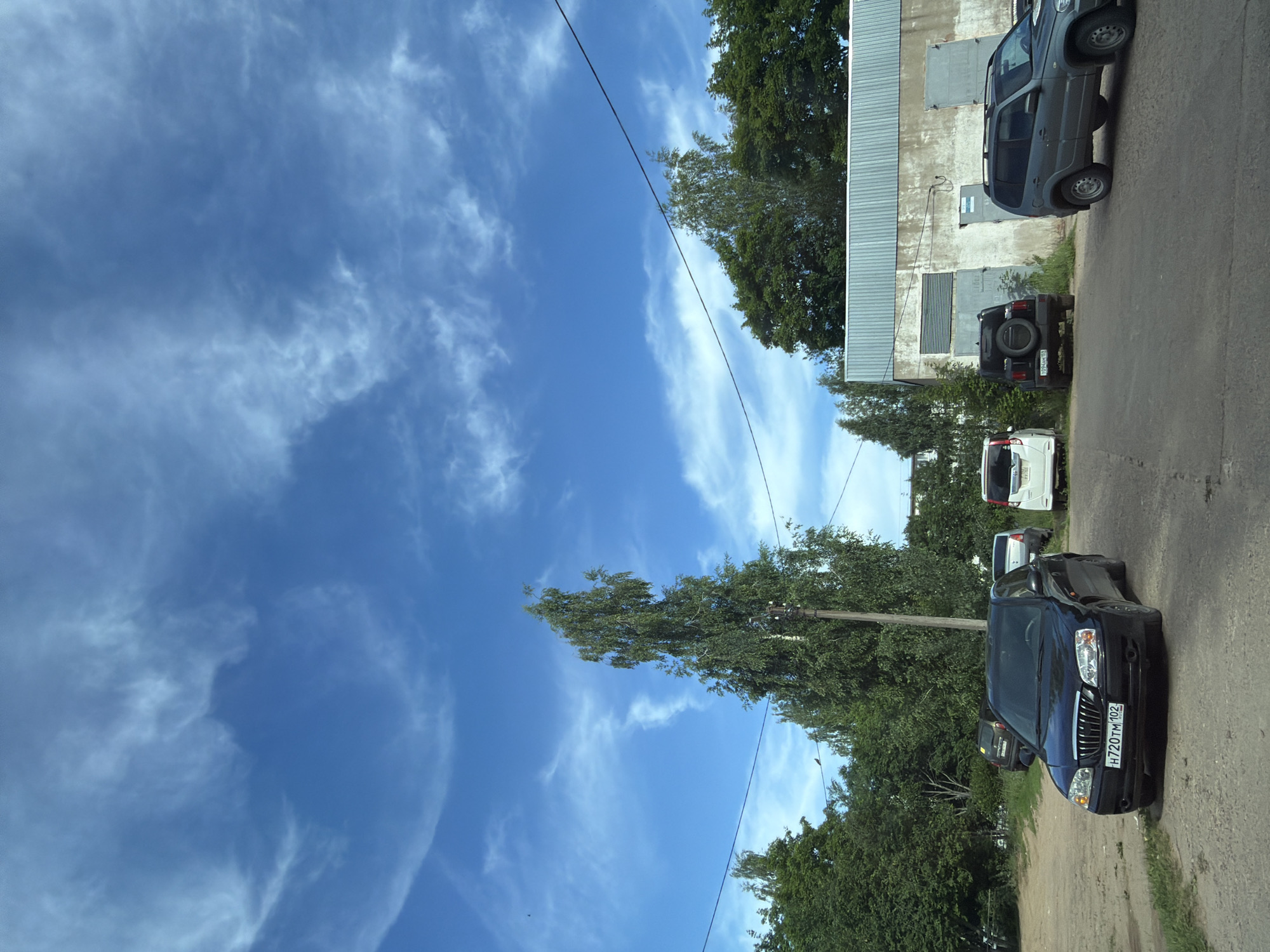 The image depicts an outdoor scene in Chaikovsky, Russia, featuring a parking lot adjacent to a concrete building with corrugated metal doors. The sky above is a vibrant blue with scattered, wispy clouds. Several vehicles are parked in the lot, including a black sedan in the foreground with a license plate reading "H720TM 102", a dark grey SUV, a black SUV with a spare tire on the back, and a white hatchback. Lush green trees and foliage surround the parking area, and power lines are visible crisscrossing the sky. A tall birch tree stands prominently in the mid-ground. The ground surface is a mix of asphalt and dirt.