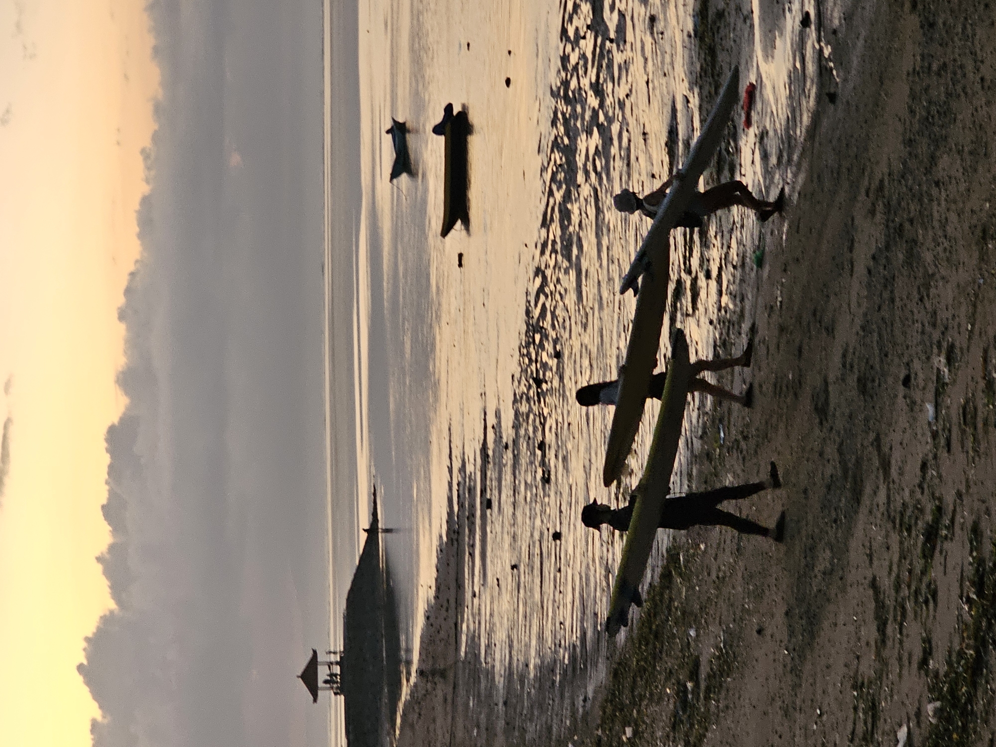 The image depicts a group of people on a beach during what appears to be sunset. The lighting suggests late afternoon or early evening, with a warm, golden glow reflecting off the water and sand. The sky is a mix of soft yellow and gray clouds, indicating partly cloudy conditions.
The main subjects are three individuals carrying surfboards towards the water. They are silhouetted against the bright background, making it difficult to discern specific features, but their posture suggests they are engaged in preparing for or finishing a surf session. The surfboards are long and narrow, typical for surfing.
In the background, there are two small boats anchored in the shallow water. Further out, a dark, pointed structure, possibly a beacon or part of a distant building, stands against the horizon. The water's surface is calm, with ripples and reflections adding texture to the scene. The beach itself is sandy, with some areas appearing wet and reflective from the receding tide. Small dark spots on the sand could be shells or debris.
The overall mood of the image is tranquil and serene, characteristic of a beach at dusk. The silhouettes and warm lighting create a peaceful atmosphere. The location context of Denpasar, Indonesia, suggests a tropical setting, which aligns with the beach environment and the presence of surfboards. No visible text is present in the image.