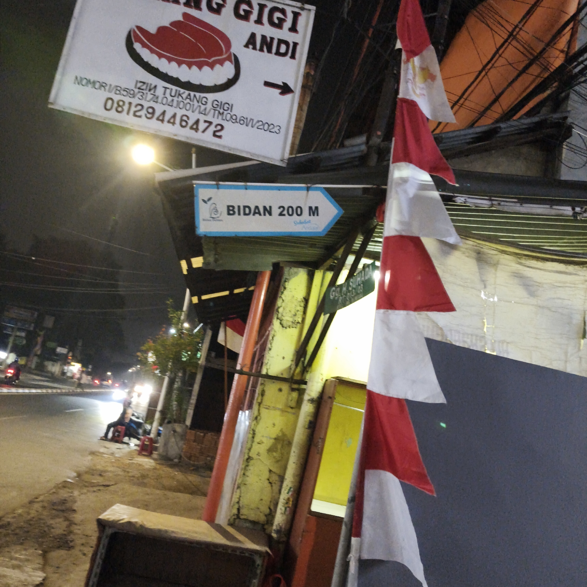 The image depicts a nighttime scene on a street in South Jakarta, Indonesia. A white sign hangs at an angle, featuring a drawing of teeth and text that reads "GIGI ANDI" and "IZIN TUKANG GIGI 08129446472". An arrow points to the right. Below this, a blue directional sign indicates "BIDAN 200 M" with an arrow to the right. Indonesian flags, red and white, are visible, one prominently in the foreground. The street is paved, with a sidewalk in the foreground where a person is seated next to a motorcycle. Traffic lights and streetlights illuminate the scene. Buildings and electrical wires are visible in the background.