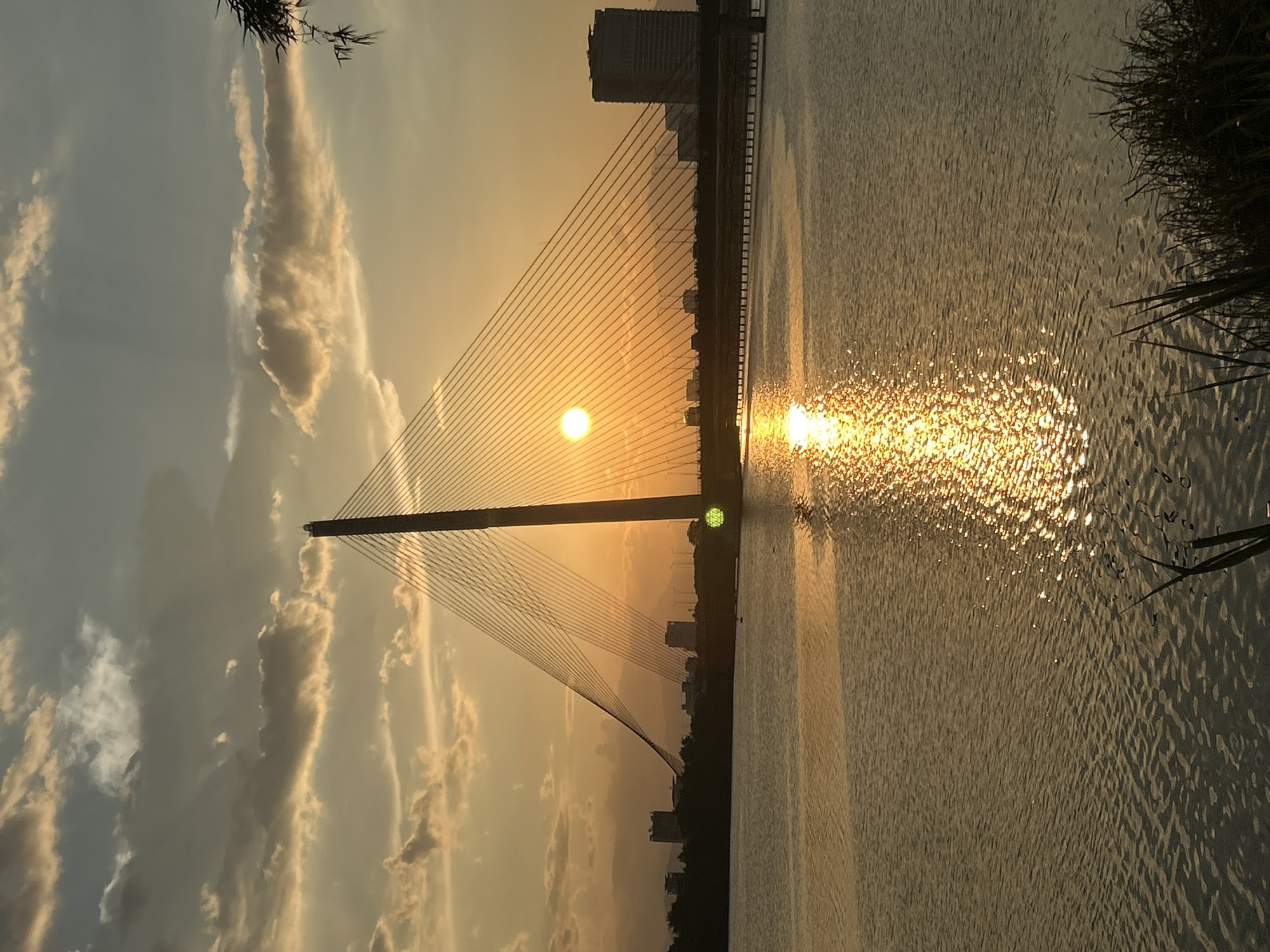 The image depicts a serene sunset over the Hàn River in Đà Nẵng, Vietnam, with the iconic Dragon Bridge prominently featured. The sun, a brilliant orb, is partially obscured by clouds, casting a warm, golden glow across the sky and reflecting on the rippling surface of the river. The cable-stayed bridge's distinctive pylons and delicate cables stretch across the frame, creating a dramatic silhouette against the vibrant sunset. In the background, the city skyline is visible, with buildings of varying heights adding a sense of depth to the scene. The tranquil water, the dramatic bridge, and the colorful sky combine to create a breathtaking panoramic view of Đà Nẵng at dusk.