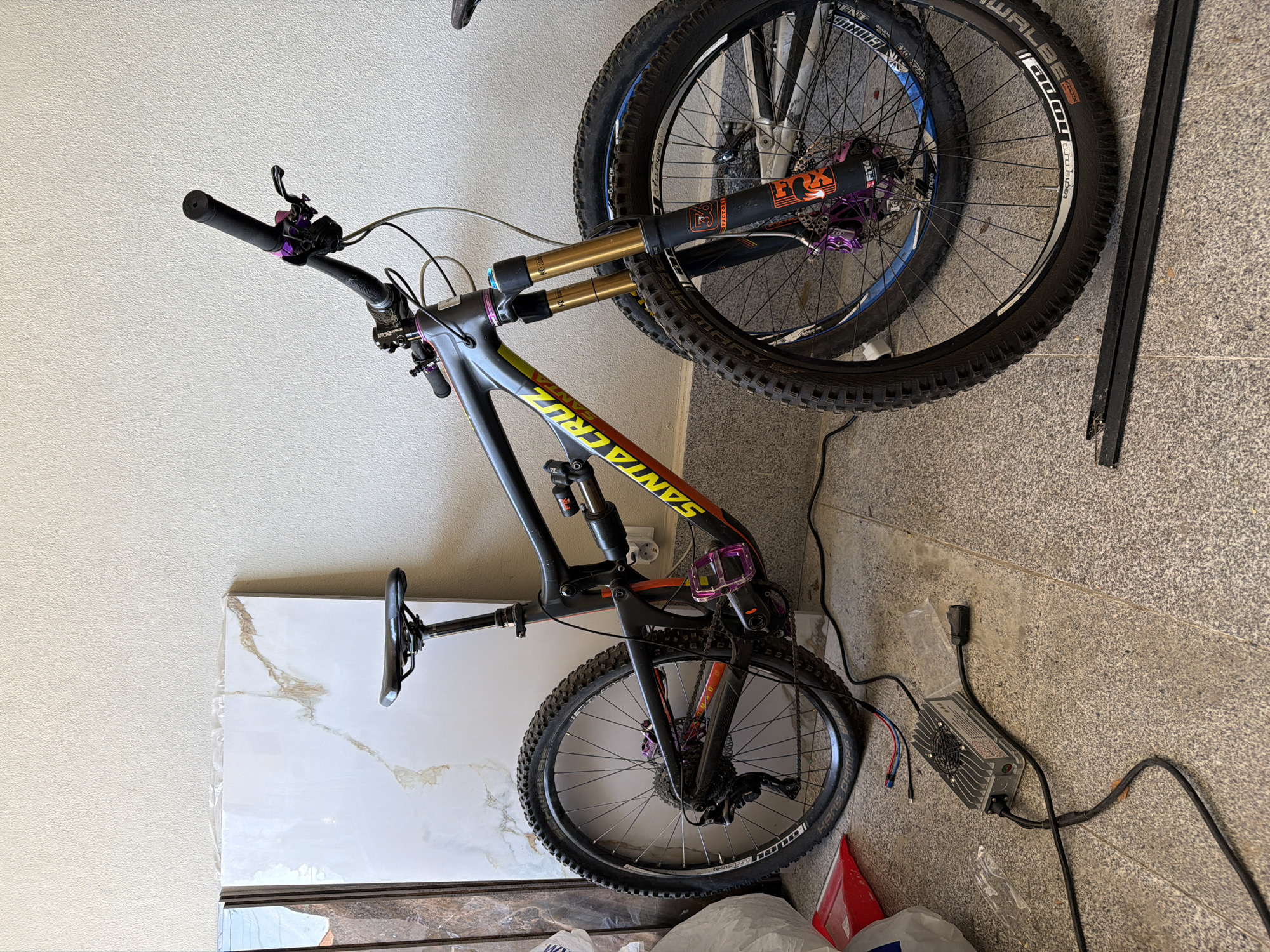 A mountain bike is leaning against a white wall in what appears to be a garage or storage area. The bike is a Santa Cruz model, with black frame, yellow and orange lettering, and purple accents on components such as the pedals and brake levers. The bike has front and rear suspension, with a gold-colored front fork. The wheels have knobby tires. An electronic device with wires and a fan, possibly a battery charger, is on the floor near the bike. A stack of tiles wrapped in plastic is next to the bike. There are other miscellaneous objects scattered around the floor, including pieces of a black strip. The location is unknown.