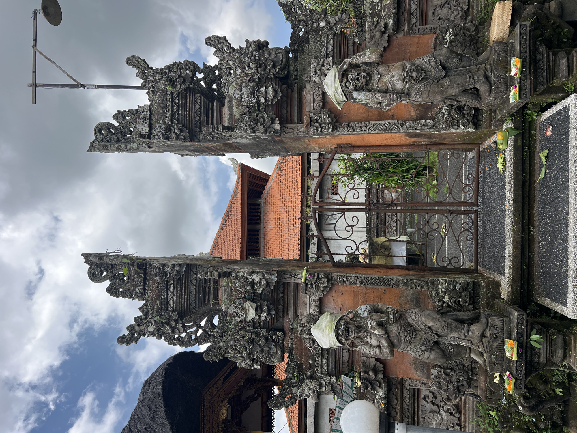 The image depicts a traditional Balinese entrance gate, likely leading to a temple or a private residence, located in Ubud District, Indonesia. The gate is made of stone and ornately carved with mythological figures and floral motifs. Two stone statues stand guard on either side of the gate, each adorned with a white cloth tied around its head. Offerings of flowers and food are placed at the base of the statues, indicating religious or spiritual significance. Behind the gate, a building with a red tile roof is visible. The sky is overcast with gray clouds, and a lamppost stands nearby. The overall scene conveys a sense of cultural heritage and religious practice.