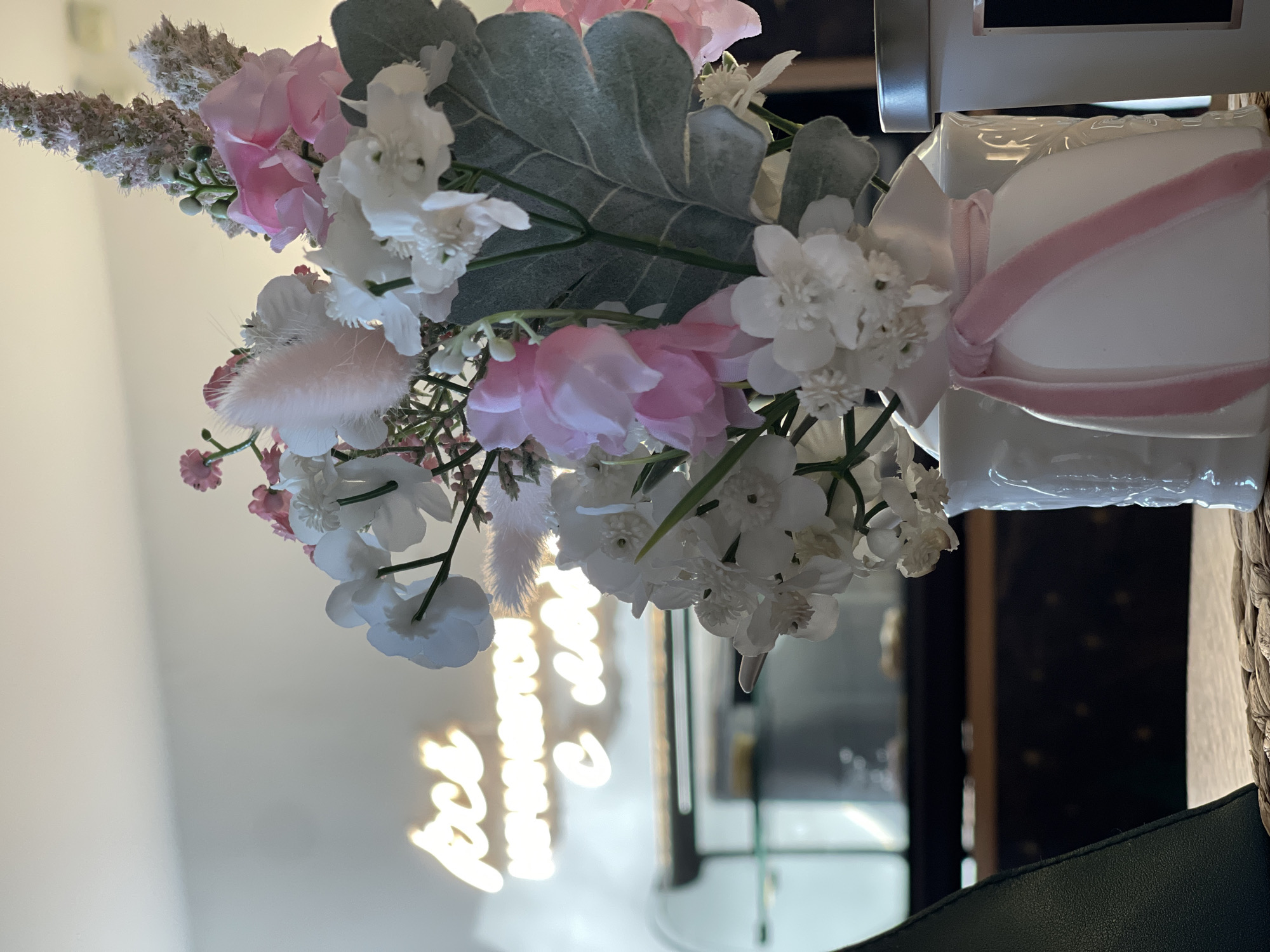 The image depicts a floral arrangement with white and pink flowers, featuring a large, velvety grey-green leaf. The arrangement is tied with a pink ribbon around a white, rectangular container. In the background, there is a blurred reflection of neon lettering, possibly indicating a shop or restaurant name, and a glimpse of what appears to be a glass shelf or display. The presence of neon lighting and the floral arrangement could suggest an interior setting of a commercial space. There is no information to indicate location in Votkinsk. There are no people depicted in the image.