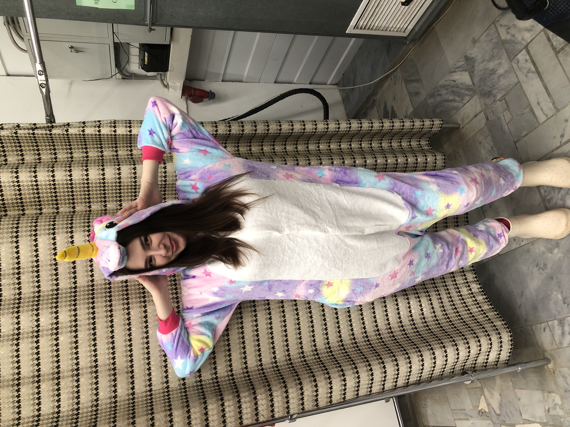 A person wearing a unicorn onesie is lying on a bed indoors. The location is Chaikovsky, Russia. The person is facing the camera and smiling. The bed has a patterned cover. The floor is tiled. There is ventilation ductwork visible near the top of the frame.