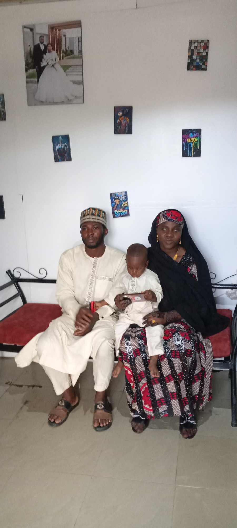 A family portrait shows a man, woman, and young child sitting on a red cushion bench with a black metal frame against a white wall. The man, dressed in an off-white long-sleeved outfit and a patterned cap, sits to the left. The woman, wearing a black headscarf and a patterned red and black skirt, sits to his right. The child is sitting on the woman's lap and using a small electronic device, possibly a tablet. Behind the group, the wall is decorated with various framed images, including a wedding photo and other artistic prints. The location is Unknown, Unknown.