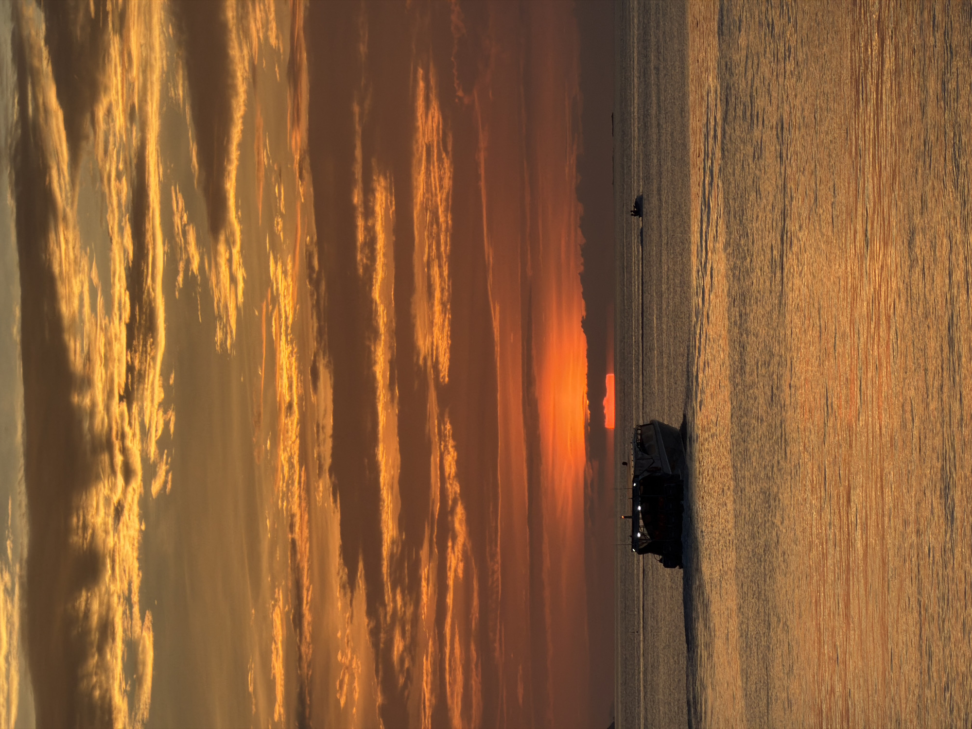 This is a landscape photograph taken in the Ko Samui District of Thailand, featuring a dramatic sunset over a body of water. The image is oriented vertically, with the sky occupying the left two-thirds and the water the right third.
The sky is filled with long, striated clouds, painted in vibrant hues of orange, yellow, and gold, indicative of a sunset. The sun itself appears as a bright, fiery orange sliver on the horizon, casting a warm glow across the scene.
The right side of the image depicts a vast expanse of water, reflecting the colors of the sunset, creating a shimmering, textured surface. A small dark object, possibly a boat or a vehicle, is visible in the distance on the water. In the foreground on the right, a dark-colored vehicle, possibly an all-terrain vehicle or similar rugged transport, is positioned on what appears to be a sandy or muddy shore. The vehicle has lights on its roof.
The overall mood of the image is serene and awe-inspiring, capturing the beauty of a tropical sunset. The time of day is clearly sunset, with the warm, golden light bathing the clouds and water. The location cues suggest a coastal or beach area in Thailand. There is no visible text in the image.