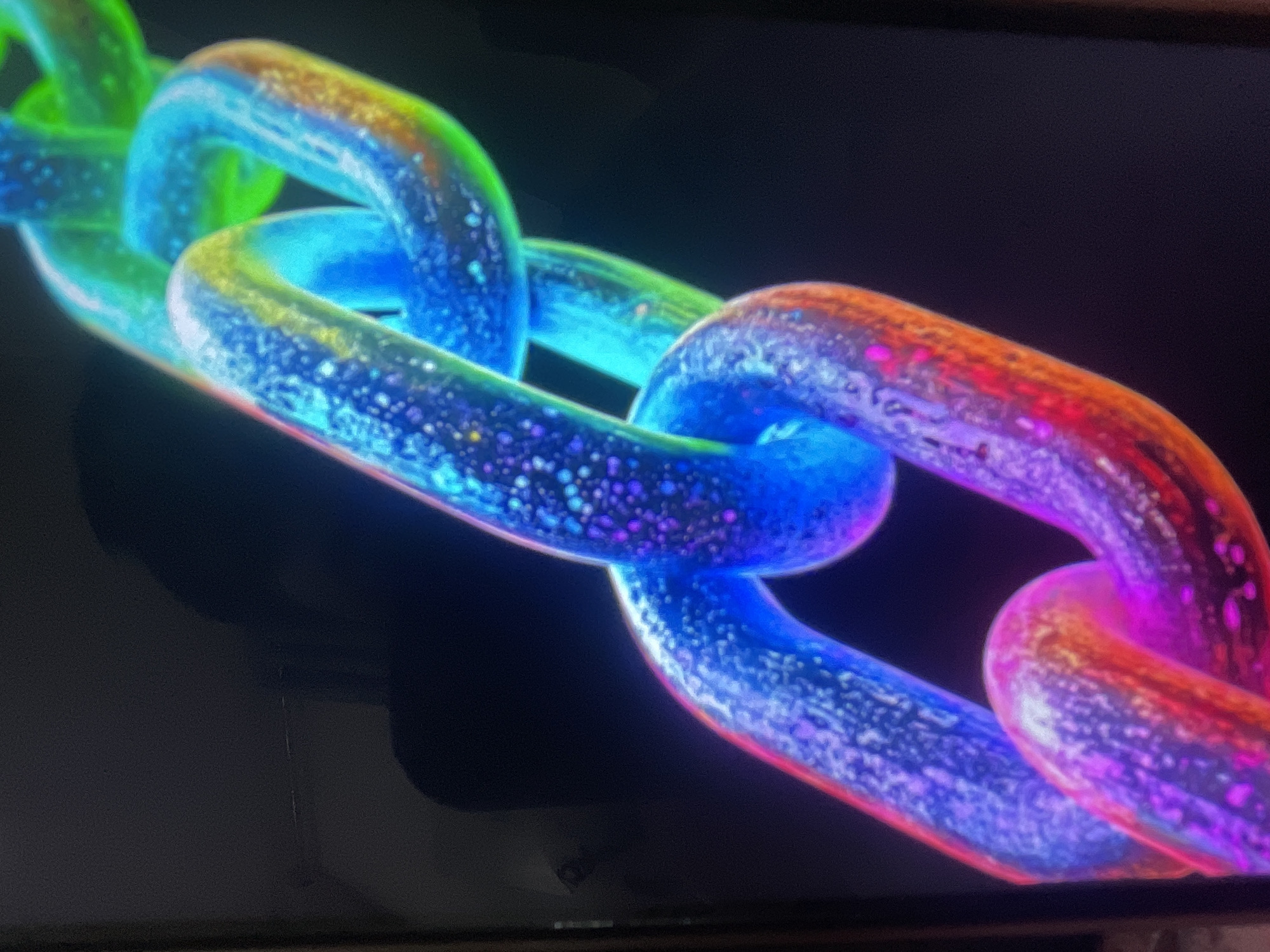 The image displays a close-up of a multi-colored chain against a dark background. The chain links are illuminated with a gradient of colors, including blue, green, yellow, orange, and purple, with a textured, speckled surface. No people, actions, interactions, or specific environmental details are discernible. The image does not contain any information that would allow for its localization in Bishkek City, Kyrgyzstan.