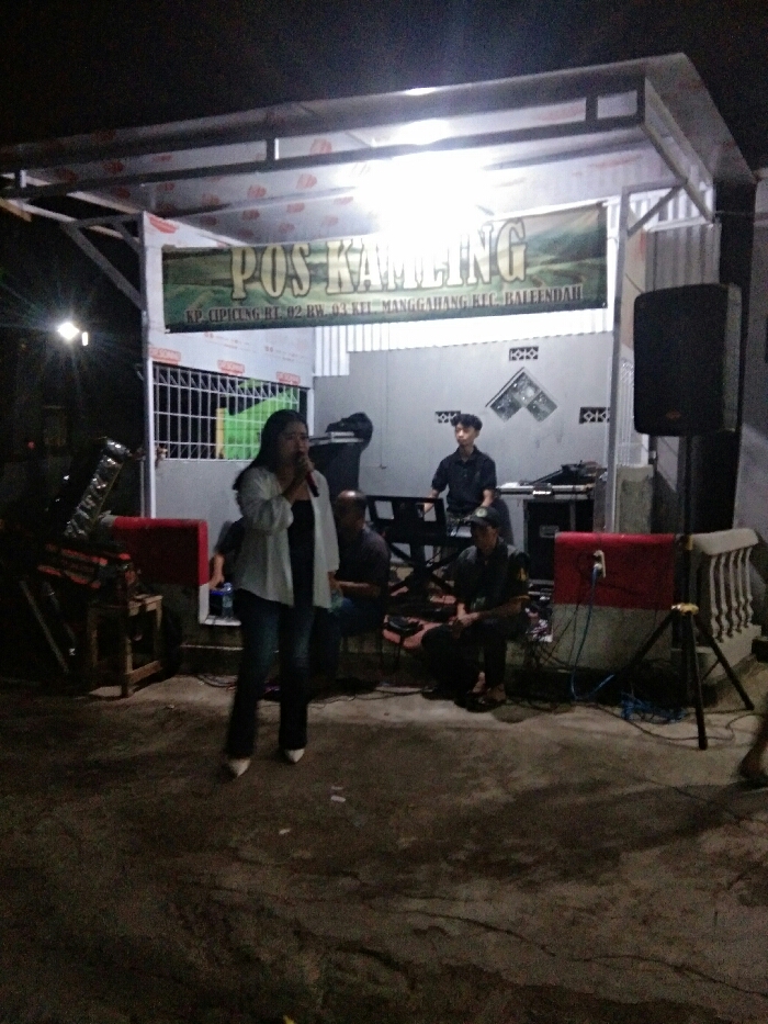 The image captures a nighttime outdoor performance or community gathering under a temporary, open-sided structure, likely a canopy, supported by metal beams. The scene is brightly lit by overhead artificial lights, contrasting with the dark surroundings.
In the center foreground, a woman stands holding a microphone to her mouth, appearing to sing or speak. She is wearing a light-colored (possibly white) long-sleeved shirt over a dark top, dark trousers, and light-colored pointed shoes.
Behind her, several men are positioned as if part of a band or accompanying musicians. To her right (viewer's left), a man is seated, partially obscured. Further back and to the right, another man is seated at a keyboard or synthesizer, seemingly playing the instrument. Another man is visible seated on the right side of the stage area. On the far left, a stand that appears to be for a drum set or other percussion instrument is visible.
The performance area is set up against a light-colored wall or building, which features some decorative or ventilation blocks in its upper section.
Above the stage, a prominent green banner hangs, displaying white text. The main text reads "POS KAMLING". Below this, in smaller white letters, it states: "KP. CIPICUNG RT. 02 RW. 03 KEL. MANGGAHANG KEC. BALEENDAH".
Equipment for the performance is visible around the stage. On the far right, a large black speaker is mounted on a stand. Closer to the ground on the right, a smaller red and white speaker or amplifier is placed, surrounded by various cables.
The ground surface in the foreground appears to be dark, possibly concrete or pavement, showing some texture. The overall atmosphere suggests a casual, local event or performance taking place during the evening.