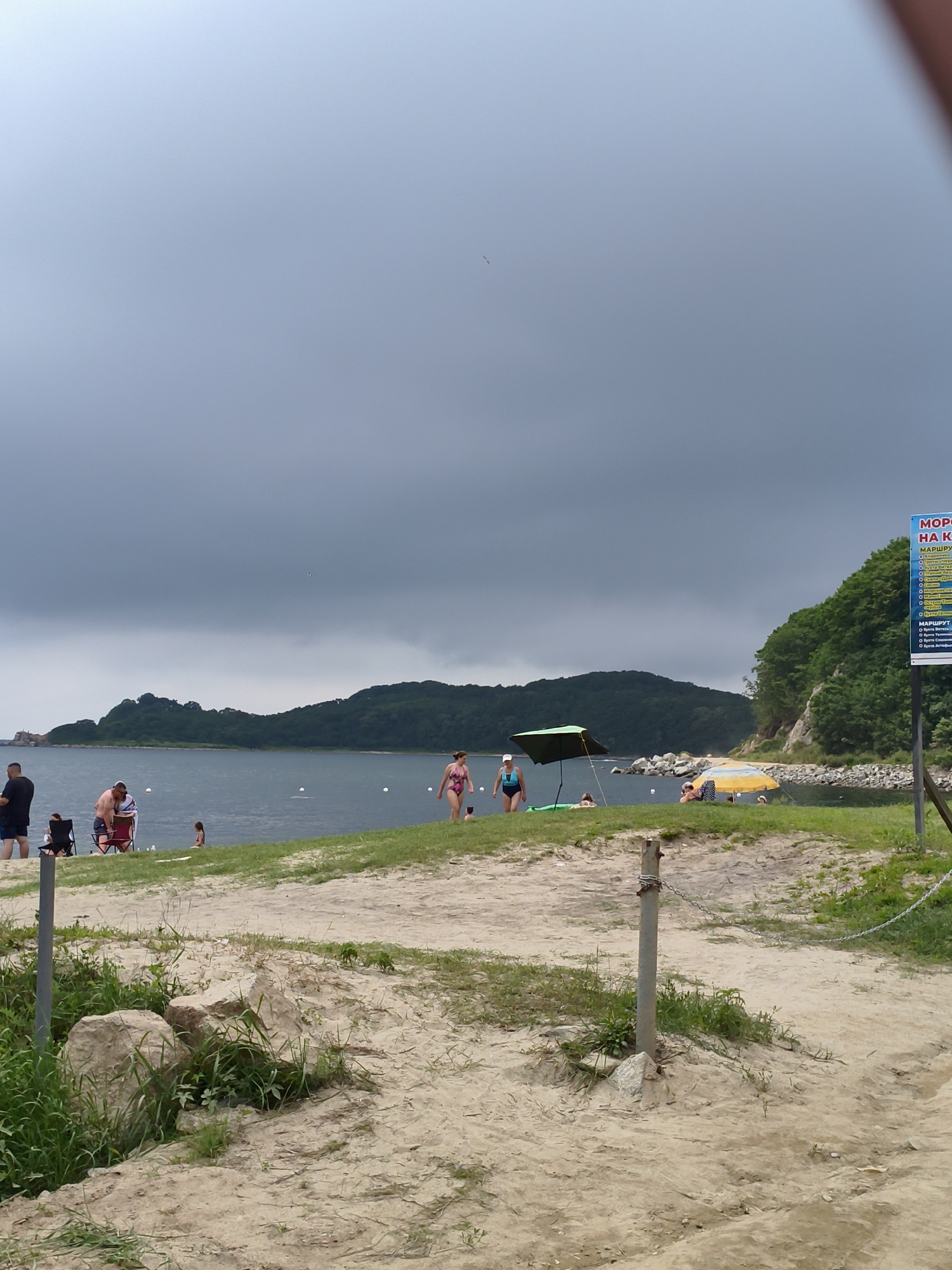 The image depicts a beach scene in Andreevka, Russia. A group of people are scattered along the sandy shore and in the shallow water, engaging in various activities. On the left, two men and a child are present, with one man standing and the other two sitting in chairs. Further to the right, a woman in a pink swimsuit walks along the water's edge. In the water, several other individuals can be seen, some swimming and others relaxing. The environment is characterized by a calm sea, a green, tree-covered hill in the background, and a cloudy sky. A signpost with text in Russian is visible on the right side of the image.