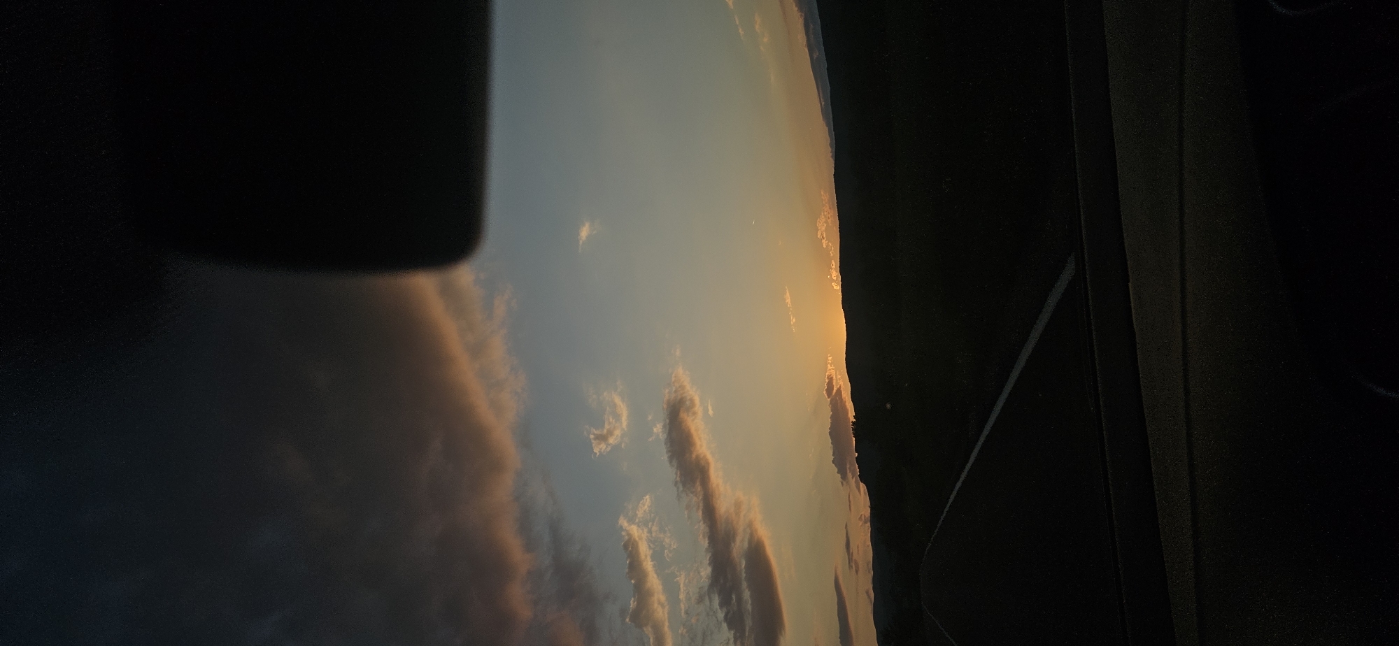 A horizontal, wide-angle photograph, taken from the interior of a vehicle, captures a sunset over a rural landscape in the vicinity of Dolna Banya, Bulgaria. The upper portion of the sky transitions from dark, heavy clouds with grey and subtle pink hues in the upper-left, to a clear, pale blue in the upper-mid, then to an intense orange and yellow glow at the horizon, indicative of a sunset. Scattered, elongated cirrus clouds, reflecting warm orange and pink light, are present across the mid-sky. A faint, slender crescent moon is visible in the pale blue section, slightly right of center.
The sun itself is positioned behind a distant, silhouetted mountain range, creating a bright orange luminosity just above the dark peaks. Below the mountain range, the mid-ground landscape is rendered in deep silhouette, suggesting dense terrain or fields. In the lower-left foreground, a dark asphalt road is visible, marked by a white dashed line receding towards the center. The immediate foreground shows the dark dashboard of the vehicle, obscuring the bottom of the frame. A large, dark, irregular shape, consistent with an automotive component (e.g., rearview mirror or sun visor), occupies the upper-right quarter of the image, partially obstructing the sky. No individuals or direct interactions are visible.