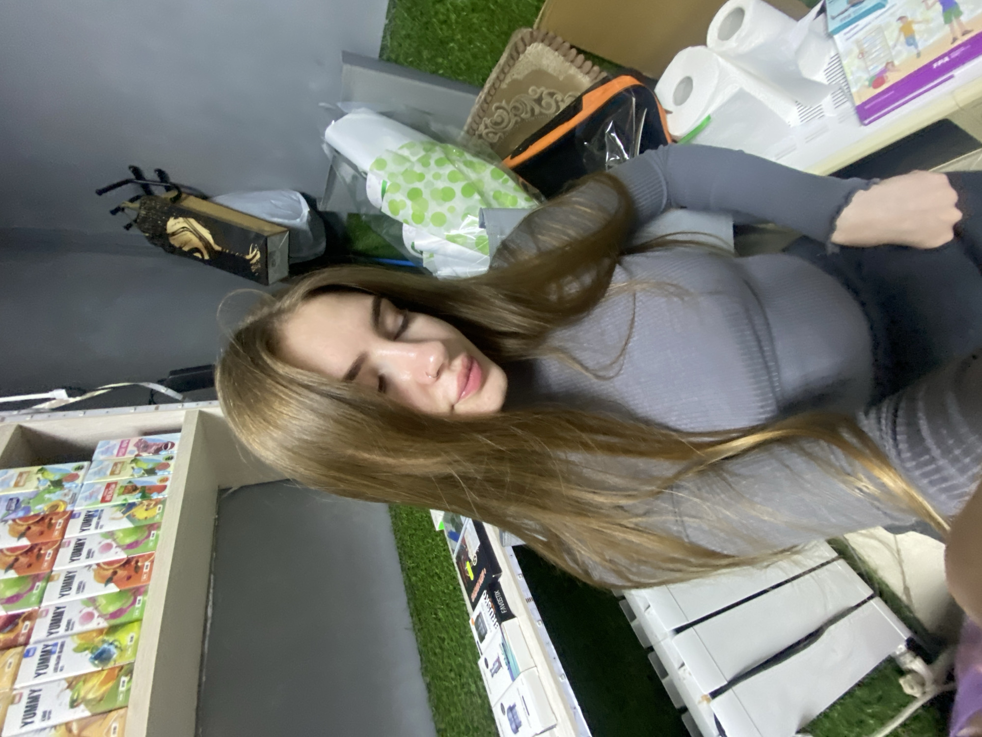 A young Caucasian female with long, brown hair is pictured in a selfie taken indoors. She is wearing a gray, long-sleeved, ribbed shirt and dark pants. Her eyes are closed, and her head is tilted slightly. The background shows a retail environment with shelves stocked with boxes of beverages labeled "YUMMY." There are also stacks of white boxes, rolls of toilet paper, a patterned cushion, and a black bag with orange trim. Artificial grass covers part of the floor and shelving units. A decorative wooden box with gold accents hangs on the wall. The location is identified as Dyakovo, Russia.