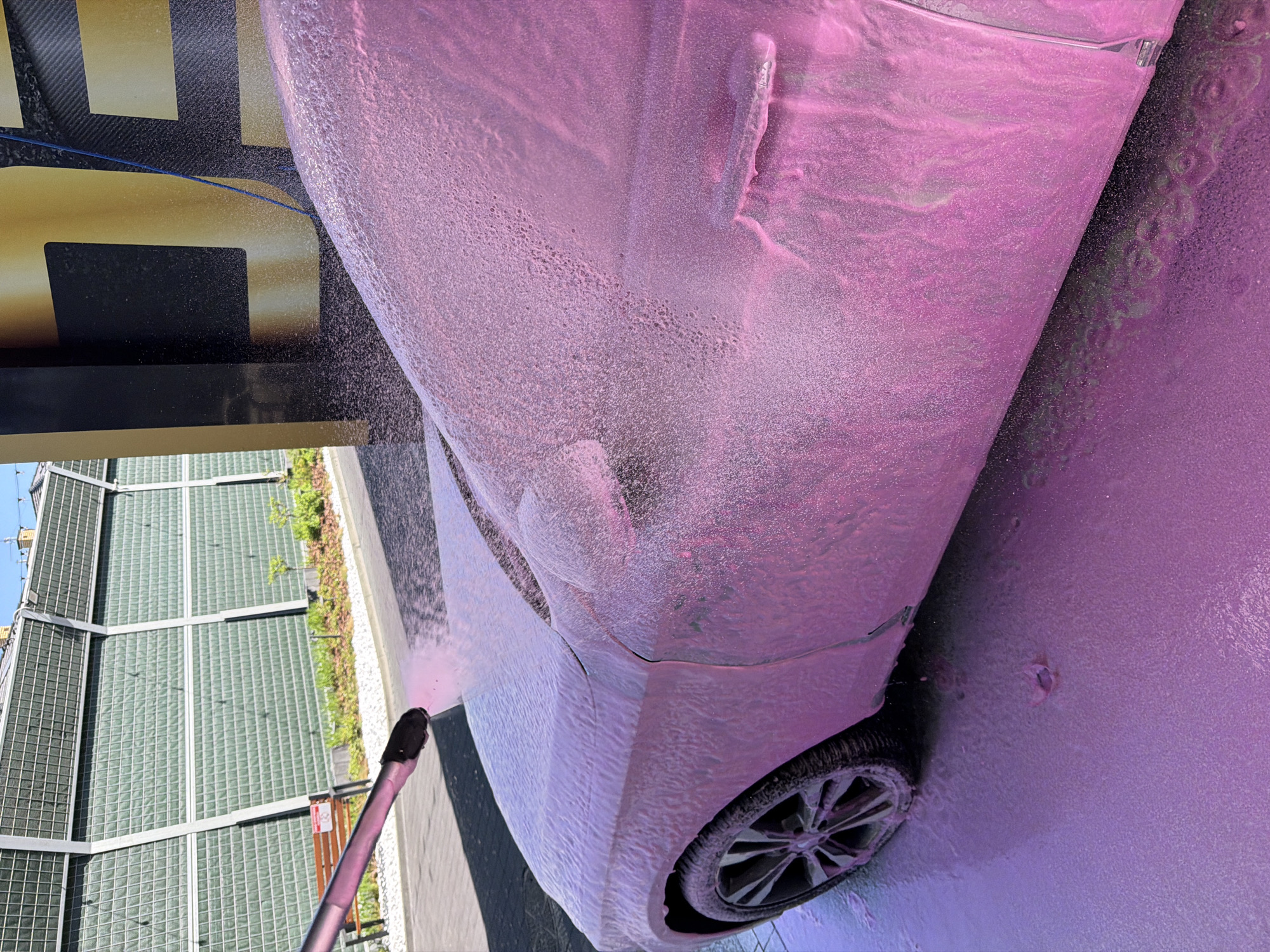 The image shows a car wash in progress. The car, likely light in color, is covered in pink foam. A pressure washer wand is visible in the lower left, directing a stream of foam onto the vehicle. In the background, a building with a glass block facade and metal framework is visible, alongside some greenery. A sign with large gold and black lettering is partially visible on the left side of the frame. The location appears to be an outdoor car wash facility in an urban or suburban setting.