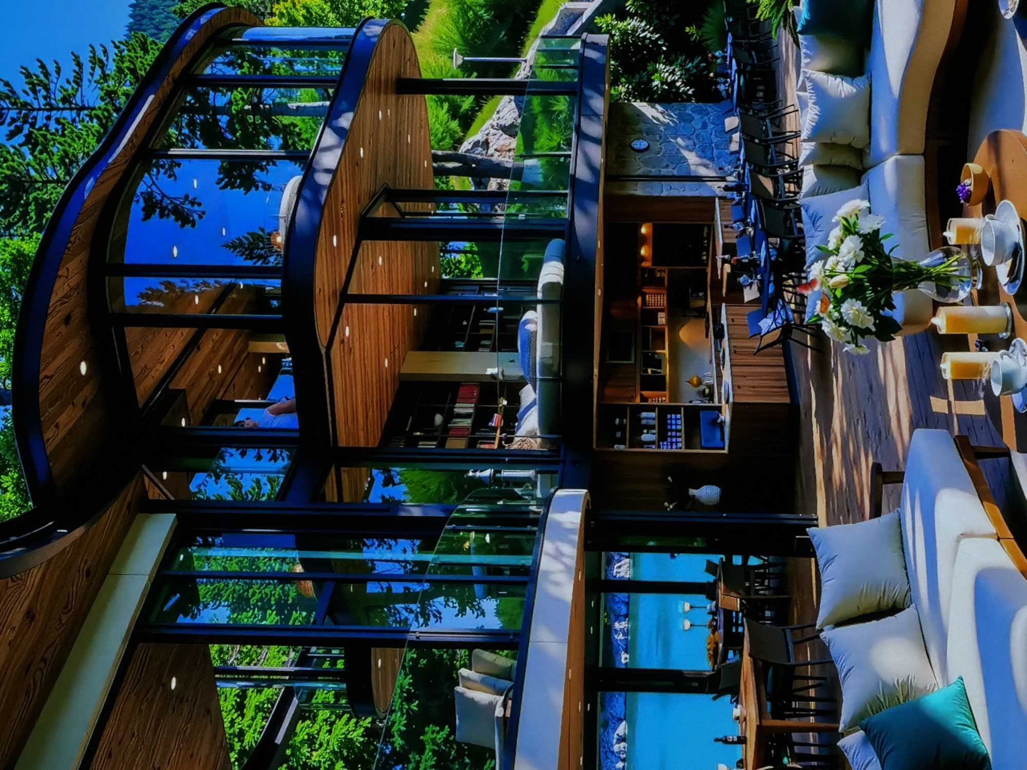 This media file captures a contemporary architectural structure nestled in a lush natural environment, likely in Sofiivska Borshchahivka, Ukraine. The multi-story building features a distinctive design with curved wooden exteriors and extensive, floor-to-ceiling glass windows that reflect a vibrant blue sky and verdant green trees.
Inside, the building showcases a warm aesthetic with wooden flooring and ceilings. Various levels are visible through the transparent walls, revealing modern shelving units stocked with bottles, decorative items, and possibly books. On one of the upper levels, a person is faintly visible, seated at a table.
Adjacent to or part of the structure is an inviting outdoor lounge area, furnished with light-colored, plush sofas and pillows. Tables are elegantly set with white cups, glasses containing orange beverages, and a prominent vase brimming with white flowers. Nearby, an outdoor dining setup with dark chairs and tables suggests a space for meals or gatherings. The surrounding landscape consists of abundant green foliage, trees, and some rocky terrain, emphasizing the building's integration with nature.
The scene is brightly illuminated by natural daylight, indicating clear and sunny weather. The overall impression is one of modern luxury, tranquility, and a seamless connection between the interior and the scenic outdoor environment. There is no visible text in the image.
