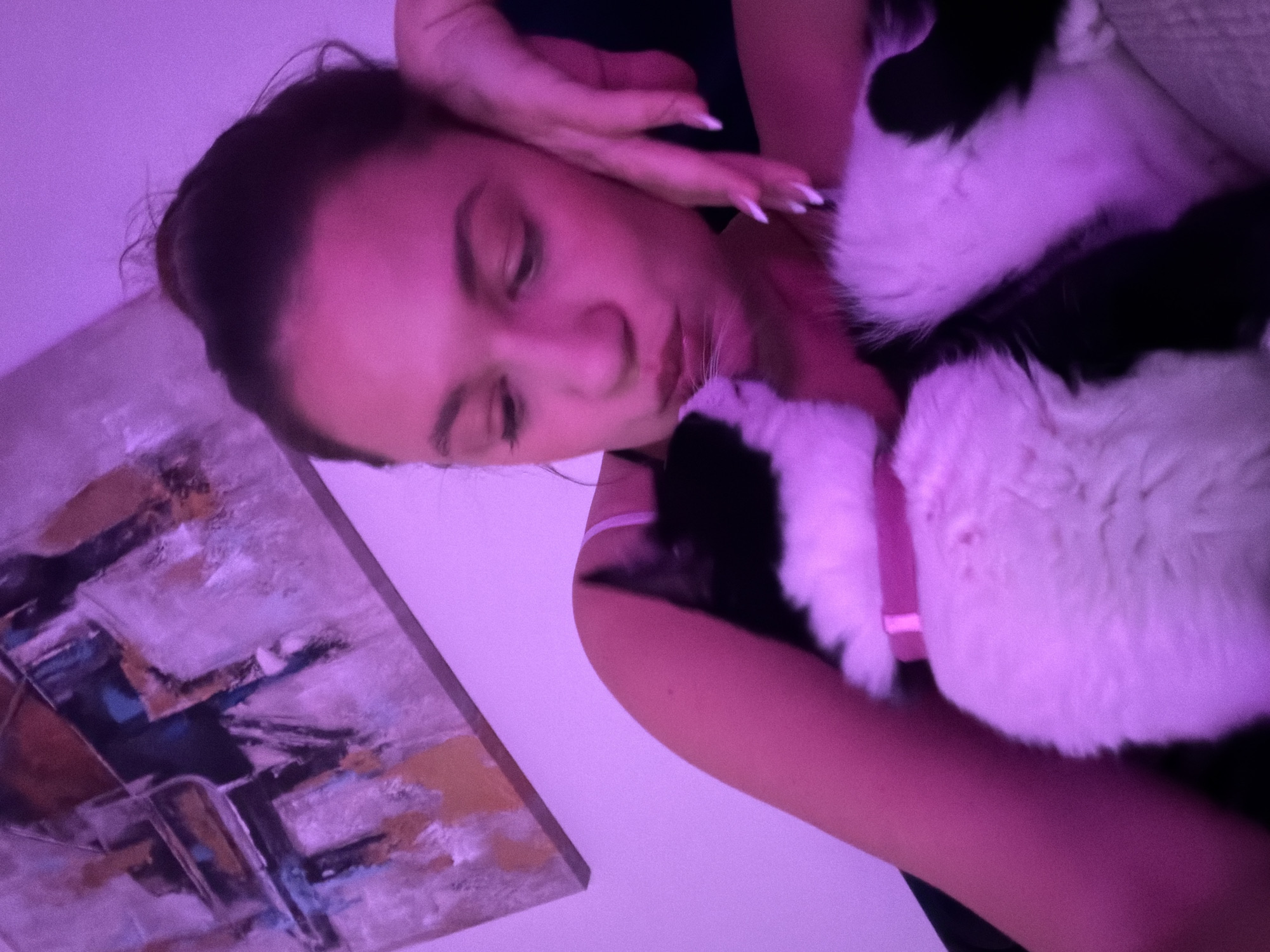 A woman is lying down with a black and white cat. The woman has her eyes closed and her hand resting on her forehead. The cat is lying on her chest. There is a painting on the wall behind the woman. The scene is lit with purple light. There is no information available to locate the scene in the city of Belgrade, Serbia.