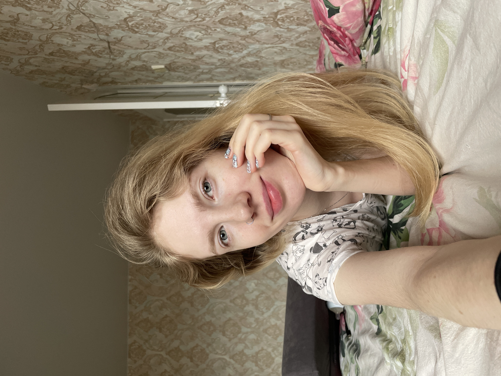 A young woman is lying on a bed in a room. The room has patterned wallpaper. The bed has a floral print comforter. The woman has long blonde hair and blue eyes. She is wearing a white shirt with a cartoon print. Her left hand is near her face. She has a nose ring. The location is inside an unidentified structure in Izhevsk, Russia.