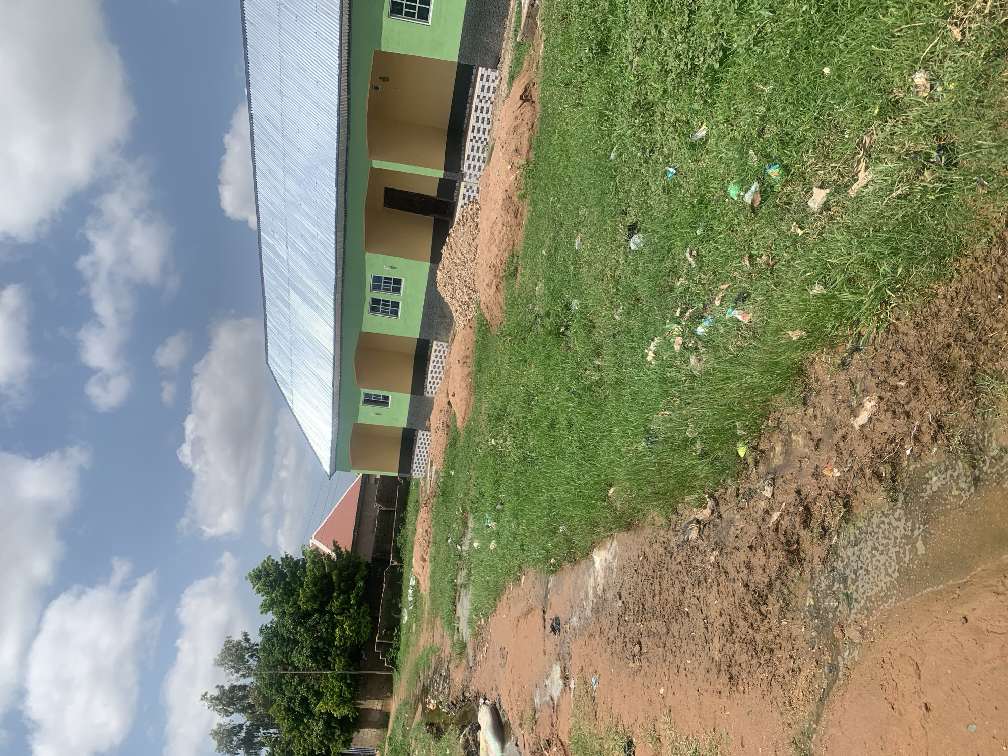 This is an exterior, eye-level, medium shot of a newly constructed building in Jalingo, Nigeria, on a partly cloudy day. The building is painted lime green and yellow with a corrugated silver roof. It has several windows with white frames and a dark brown door. In front of the building, there are piles of sand and gravel, suggesting ongoing construction. The ground is a mix of red dirt and green grass, with some areas of standing water and scattered litter. To the left of the building, a dirt path leads away, also showing signs of mud and water. Trees and other vegetation are visible in the background, along with another building with a red roof. There are no people visible in the image.