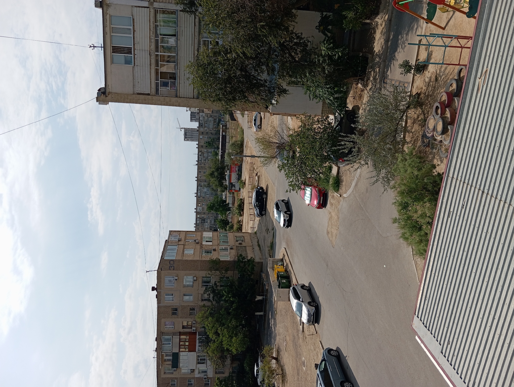 A daytime, slightly overcast scene depicts a residential courtyard in Aktau, Kazakhstan. The view is from a slightly elevated perspective, looking down onto a street and surrounding buildings. Two large, Soviet-era apartment buildings dominate the frame, one on the left and a taller one on the right. The building on the left is light brown with multiple balconies and window units. The building on the right is a pale beige and grey, also with numerous windows and visible air conditioning units.
Several vehicles are present in the courtyard: a silver SUV, a red sedan, a black SUV, and a light blue minivan. Trash bins, one green and one yellow, are visible near the entrance of a building. Trees and sparse vegetation dot the sandy and paved areas. In the lower right corner, a small children's playground with a red and blue frame and colorful seats is visible, along with a pile of old tires. In the background, other residential buildings and construction cranes suggest further development. The sky is a pale blue with scattered white clouds.