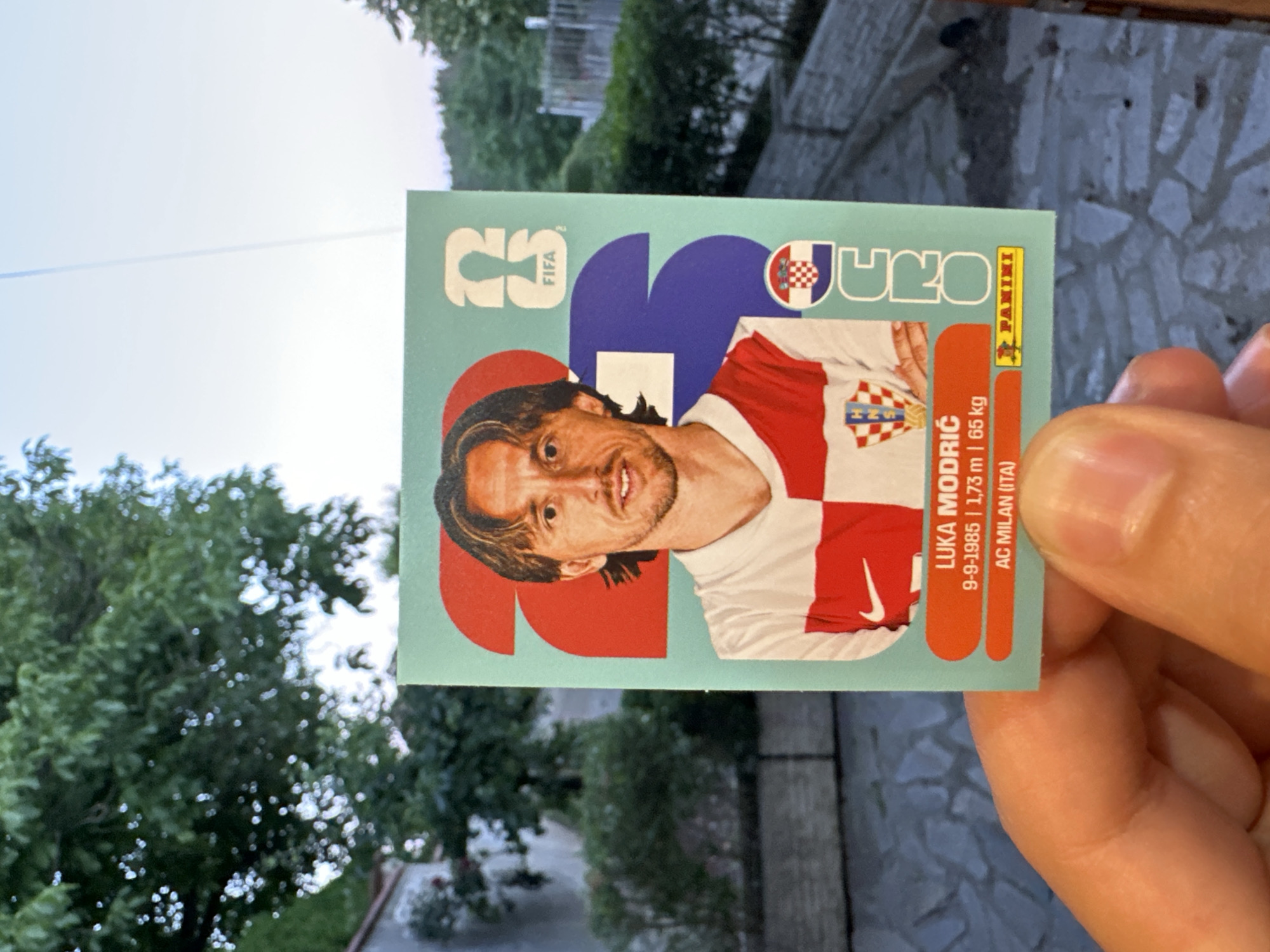 A hand holds a collectible trading card featuring soccer player Luka Modric. The card has a light blue background with a stylized image of Modric in his Croatian national team jersey. Text on the card includes his name, birthdate, height, weight, and club (AC Milan). The card also features the FIFA logo and the Panini logo. In the background, there is greenery and a hint of a building, suggesting an outdoor setting. The location is identified as the city of Prisovo, Bulgaria.