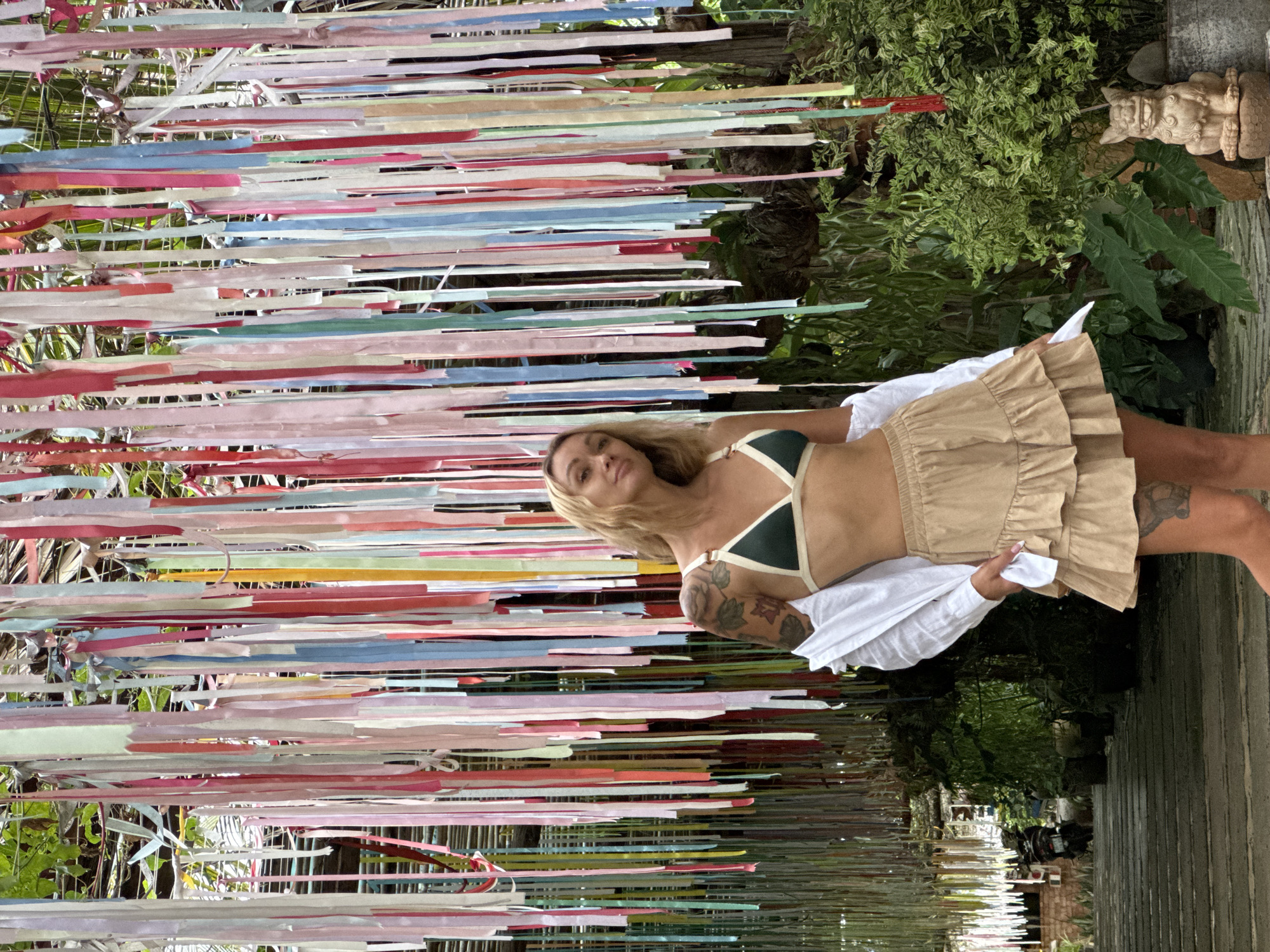 A medium shot shows a young woman lying on her right side next to a display of brightly colored ribbons. The woman has shoulder-length blond hair and a tan. She wears a dark green bikini top with white straps, a ruffled khaki-colored skirt, and a white shirt. The ribbons are stretched horizontally and are blue, pink, red, white, and gold. They are mounted on what appears to be a wire rack. In the background are green plants, and a decorative stone sculpture can be seen to the right. A wall runs along the right side of the frame. The location is Mũi Né, Vietnam.