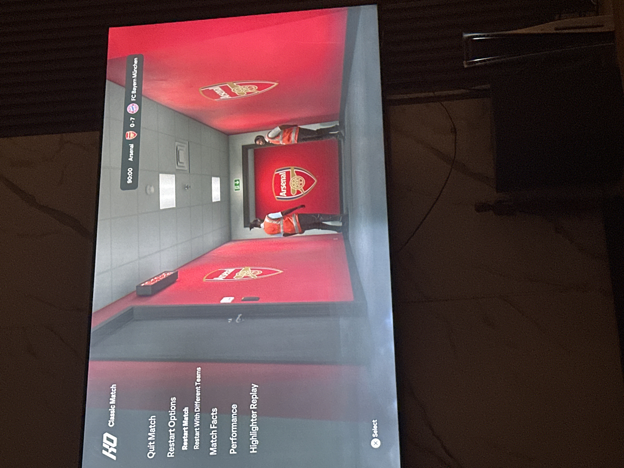 The image shows a screenshot of a video game, likely a football (soccer) game, displayed on a television screen. The game appears to be paused or in a menu screen, with options such as "Quit Match," "Restart Options," "Match Facts," and "Highlighter Replay" visible on the left side of the screen. The game's score is displayed at the top, showing Arsenal losing to FC Bayern München 0-7 in the 90th minute. The background of the game screen features a red hallway with Arsenal's team logo prominently displayed on the wall and the door. Two individuals, possibly stadium staff or security personnel wearing orange vests, are visible in the hallway.
The television screen is situated in a dimly lit room, with the overall ambiance suggesting a home entertainment setup. The presence of a person's silhouette in the background, along with some furniture and electronic devices, further supports this interpretation. There is no indication of this scene being located in Gwarinpa, Nigeria; the context provided is purely speculative. The image is a static capture of a digital display, not a photograph of an outdoor scene or a specific urban location.