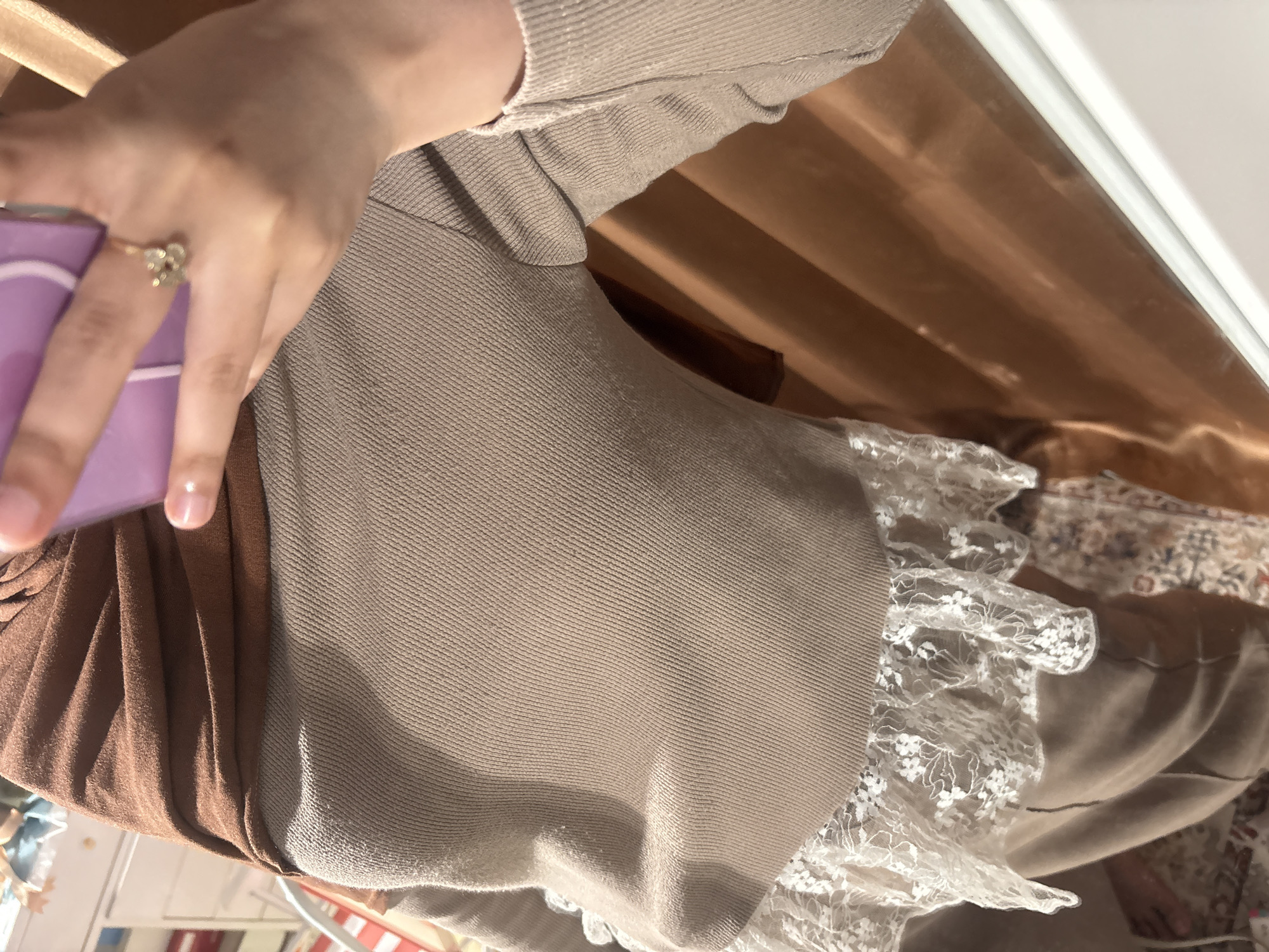 A person is holding a lavender-colored phone in their left hand, which has a gold ring on one of the fingers. The person is wearing a beige-colored sweater or cardigan with a brown dress underneath, which has a lace trim. The background appears to be a tan-colored drape. The location is unknown, and there is no information to suggest that the image was taken in Gadingrejo, Indonesia.