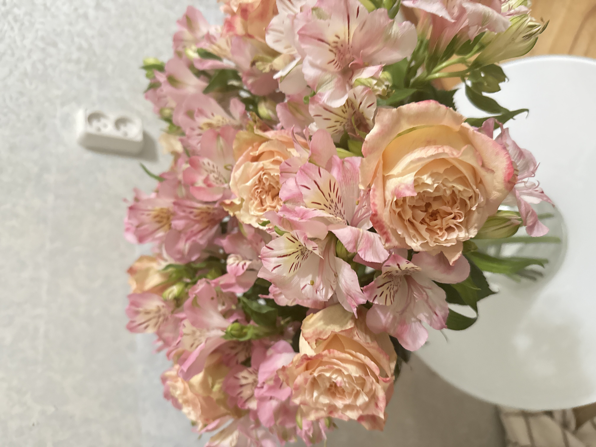 A mixed bouquet of flowers is prominently displayed, filling the majority of the frame. The bouquet consists of multiple peachy-orange garden roses with ruffled petals and numerous pink alstroemeria flowers, characterized by distinct striped markings on their inner petals. Green foliage is interspersed throughout the arrangement. The flowers are contained within a clear glass vase, which rests on a reflective white, possibly circular, table surface. The background features a light-toned, textured wall. A white double electrical outlet, consistent with European Type F (Schuko) standards, is mounted on the upper right portion of the wall. No people, animals, or explicit actions are depicted. The scene is stated to be located in Saint Petersburg, Russia.