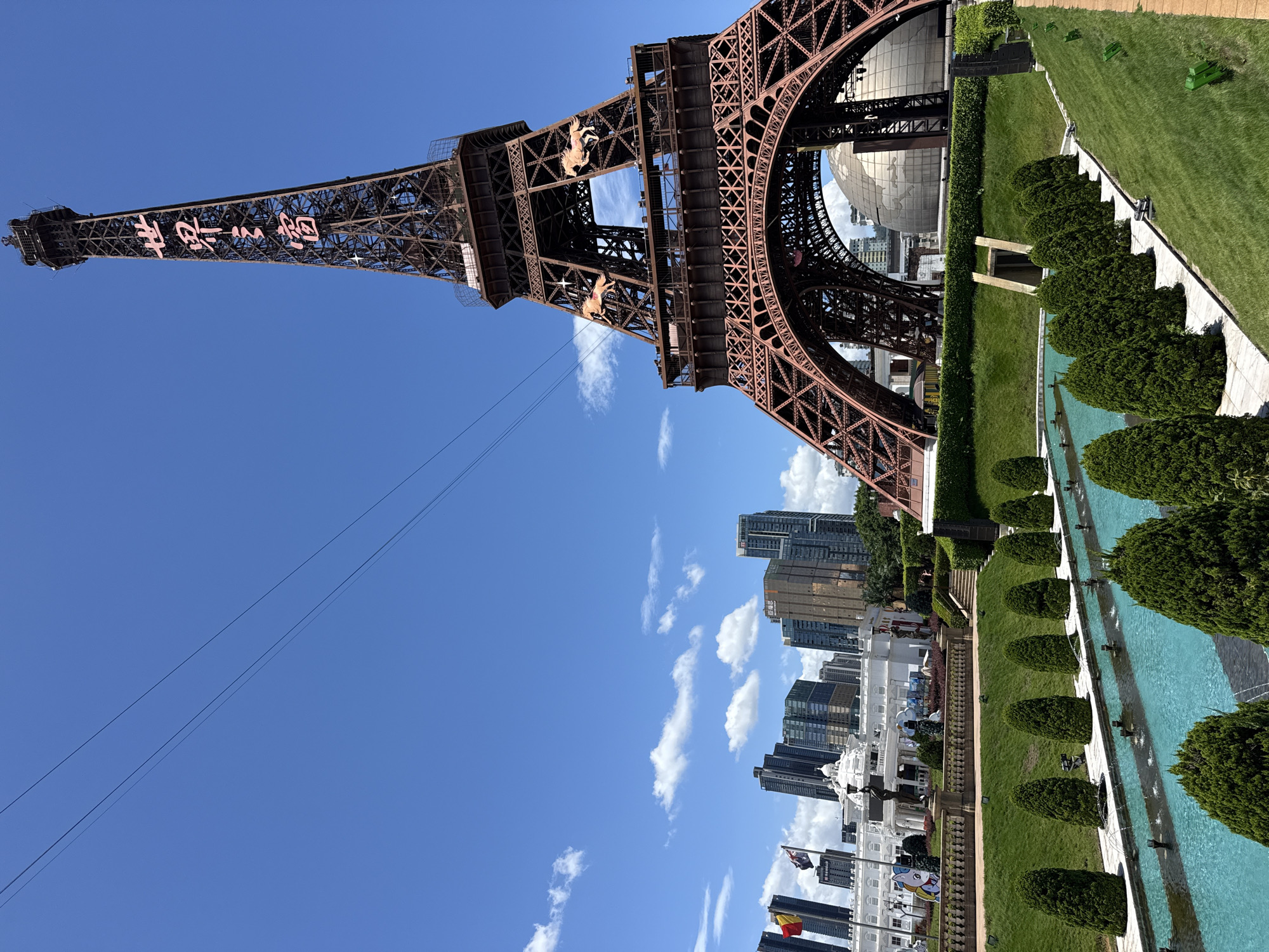 This is a medium shot of a replica Eiffel Tower in the city of Nanshan District, China, on a bright, sunny day with a clear blue sky and scattered white clouds. The tower, made of dark metal and adorned with pink Chinese characters meaning "Window of the World," is the dominant subject. Two horse statues are visible on the tower's upper levels. In front of the tower, a long, rectangular turquoise pool with a fountain system is lined with evenly spaced, conical green trees. Beyond the pool, a neatly manicured green lawn slopes upwards towards the tower. To the left of the tower, a white building with a dome and columns is partially visible, along with several tall modern buildings in the background, suggesting an urban setting. Several flags are also visible flying in front of the white building. The overall scene depicts a tourist attraction or theme park.