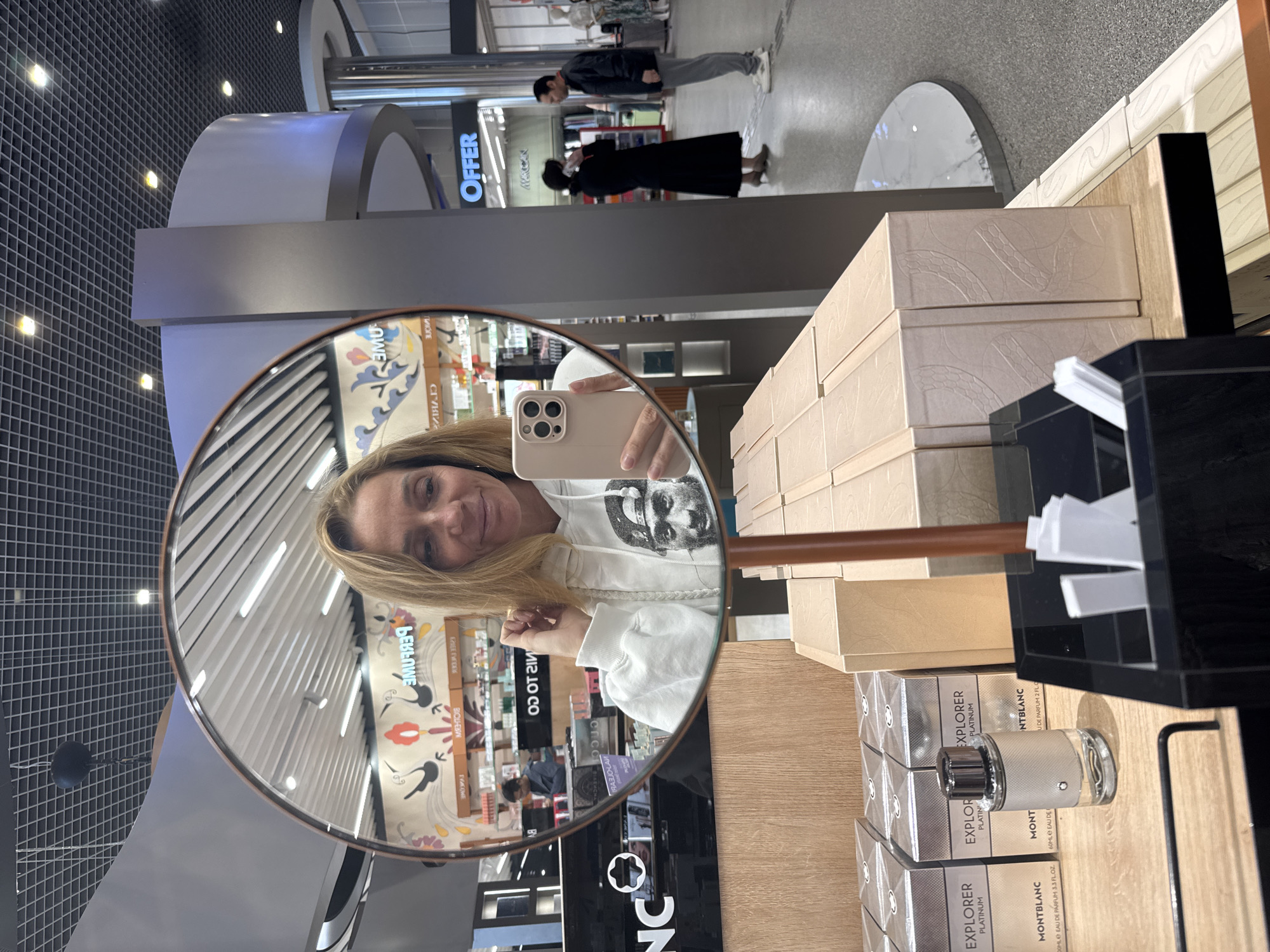 The image shows a woman taking a selfie in a round mirror, with a display of Montblanc Explorer Platinum perfumes in the foreground.
People: The woman is blonde and wearing a white hoodie with a portrait print on it. Her left hand is partially raised towards her face. There are other people reflected in the mirror, including a couple: one in a black dress and the other in a gray outfit, and additional customers and shop assistants visible in the background.
Subjects: The main subject is the woman taking a selfie. Secondary subjects include the perfumes and the people in the reflection.
Objects: The primary objects are the round mirror with a brown frame, a light pink mobile phone, the Montblanc Explorer Platinum perfume bottles and boxes, a black display stand with white perfume sample sticks, and the ceiling and wall decor of a shop.
Actions: The woman is taking a selfie in the mirror. People in the background are browsing the shop, possibly shopping.
Interactions: The woman is interacting with her phone to take a picture of herself in the mirror. Customers are interacting with the shop's products.
Environment: The setting appears to be a store, likely a perfume shop or department store, as indicated by the perfume display. The mirror provides a reflection of the shop's interior, including the wall decor with colorful designs and reversed text, the lighting fixtures, and other customers. A sign saying "OFFER" is also visible in the mirror. The location can be identified as Tashkent, Uzbekistan, based on the store name Эзилэ Фредерик, which is rendered in Cyrillic script.