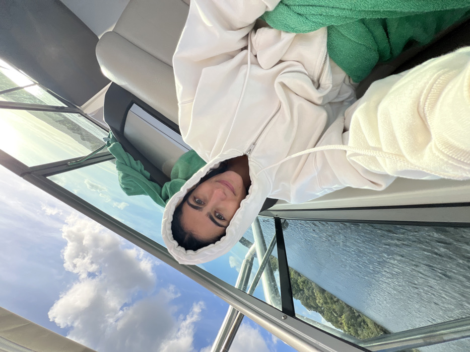 The image displays a female individual taking a selfie inside a boat on a body of water, specifically identified as the Minho River near Vila Nova de Cerveira, Portugal. The subject, with dark hair and light eyes, is positioned centrally, looking directly at the camera with a subtle smile. She is wearing a light-colored, possibly off-white, hooded zipper sweatshirt with the hood raised. Her left arm is extended towards the camera. To her immediate right, a light-colored upholstered boat seat is visible, along with a bright green towel or fabric draped over a surface. The boat's structure, including a glass windshield and metallic framing, is prominent around her. Beyond the boat's interior, a partially cloudy blue sky is visible above. The water outside the boat is calm with visible ripples, reflecting the sunlight. Distant, tree-covered hills line the horizon across the water.