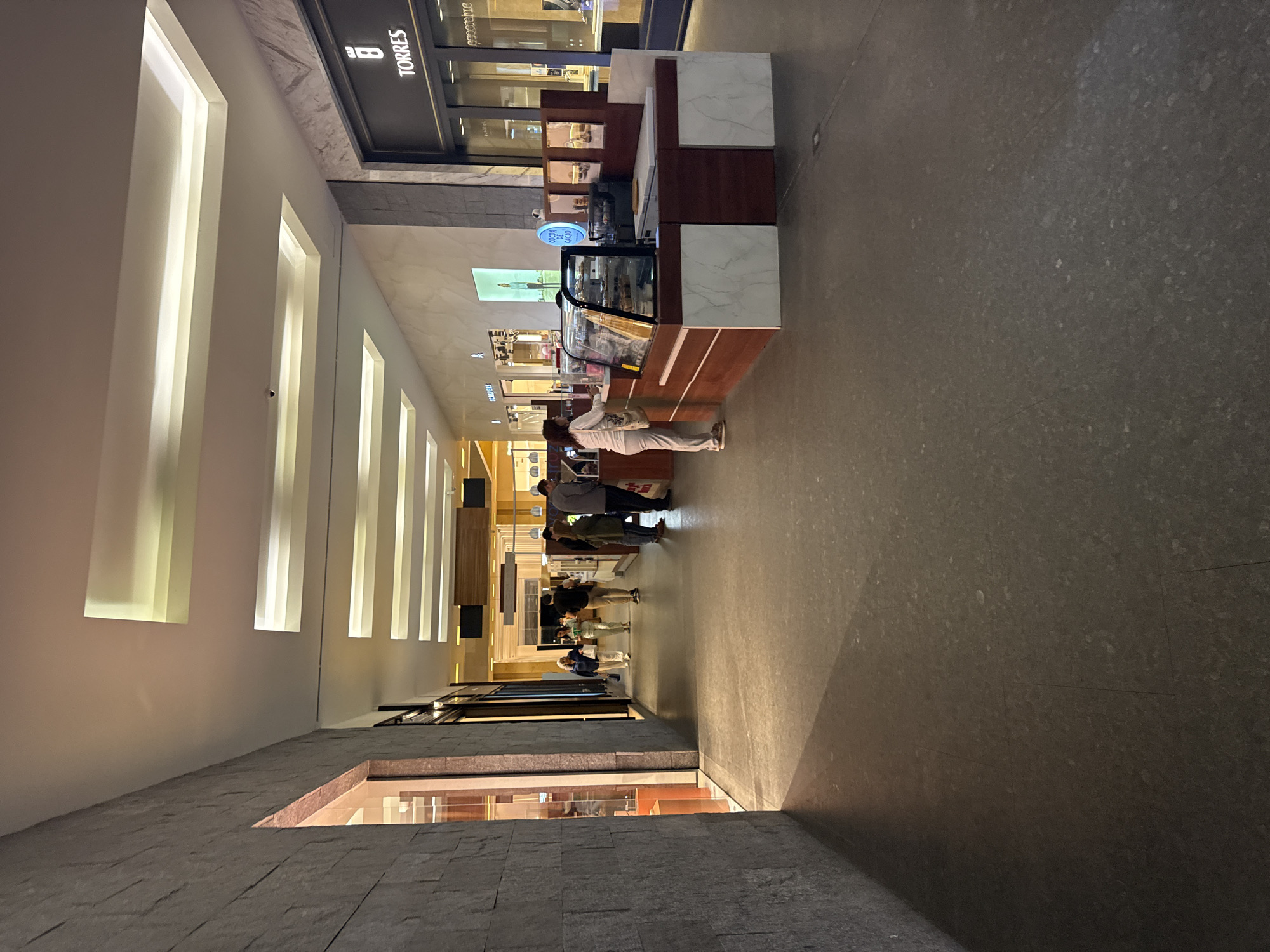 The image captures an indoor commercial corridor, likely within a mall or upscale market in Puebla, Mexico. The setting features a long, well-lit passageway with textured dark gray flooring on the right and light-colored walls with vertical illuminated panels on the left.
In the mid-ground, on the right side of the corridor, is a contemporary food or beverage kiosk. This counter features a dark wooden base with elegant white marble-patterned tops and accents. A glass display case on the counter holds various pastries or desserts. Behind the counter, a vendor is partially visible, serving customers. Several people are gathered around the kiosk, with one individual in white attire standing directly at the counter, appearing to make a purchase or order. Further down the corridor, more individuals can be seen walking or congregating, suggesting a moderately busy environment.
Behind the kiosk, a store facade with dark paneling is visible, displaying various items in its window.
Notable details include the artificial lighting that illuminates the entire space, suggesting either nighttime or an interior area without natural light. The architecture of the walls with recessed, glowing panels contributes to a modern and sophisticated ambiance.
Visible text includes "TORRES" on a dark sign above the store behind the kiosk. A blue circular sign on the kiosk counter reads "COCINA DE CACAO," suggesting a focus on chocolate-based products. Partially visible further down the corridor, another sign appears to read "CHOCOLATES."
