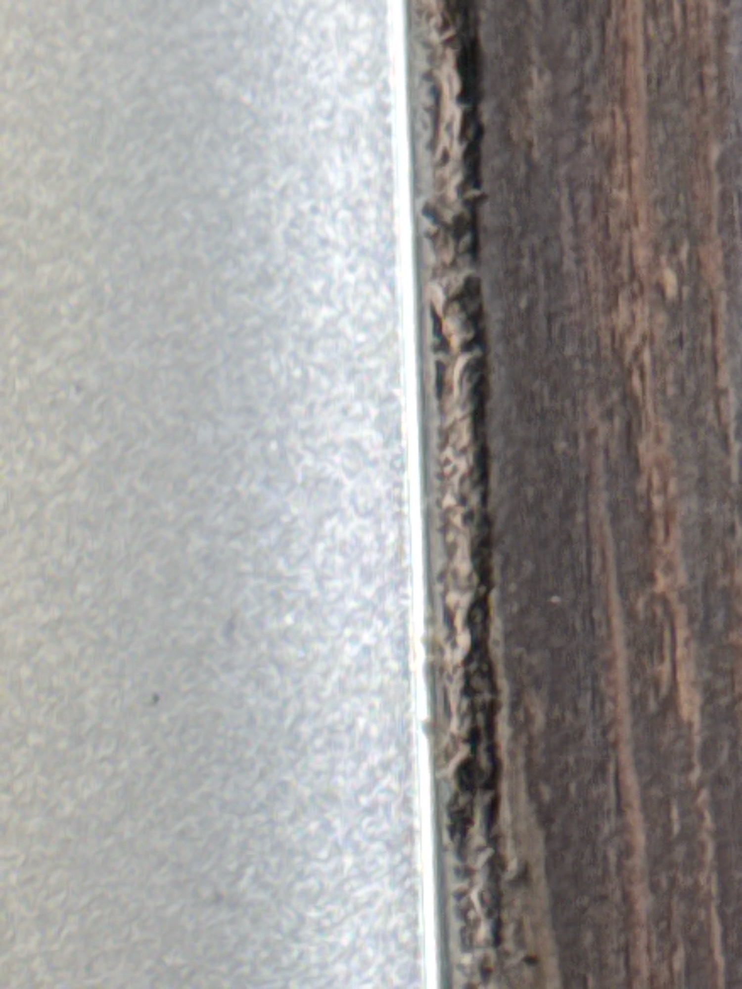 The image is a close-up, top-down view of a textured surface, possibly a wall or floor, showing a distinct border between two materials. On the left, a light-colored, textured surface with a metallic sheen fills approximately half the frame. The texture appears to be a repeating pattern of small, irregular shapes. On the right, a darker, rougher surface with a wood-grain pattern dominates the other half. The wood grain runs vertically and exhibits variations in color, ranging from dark brown to lighter reddish-browns, with visible imperfections and splintering along the edge. The two surfaces meet at a sharp, vertical line, where some debris or sealant is visible along the edge of the wood grain. The lighting appears to be diffused, casting no strong shadows. The scene is static, with no people or overt actions depicted. The location is unknown.
