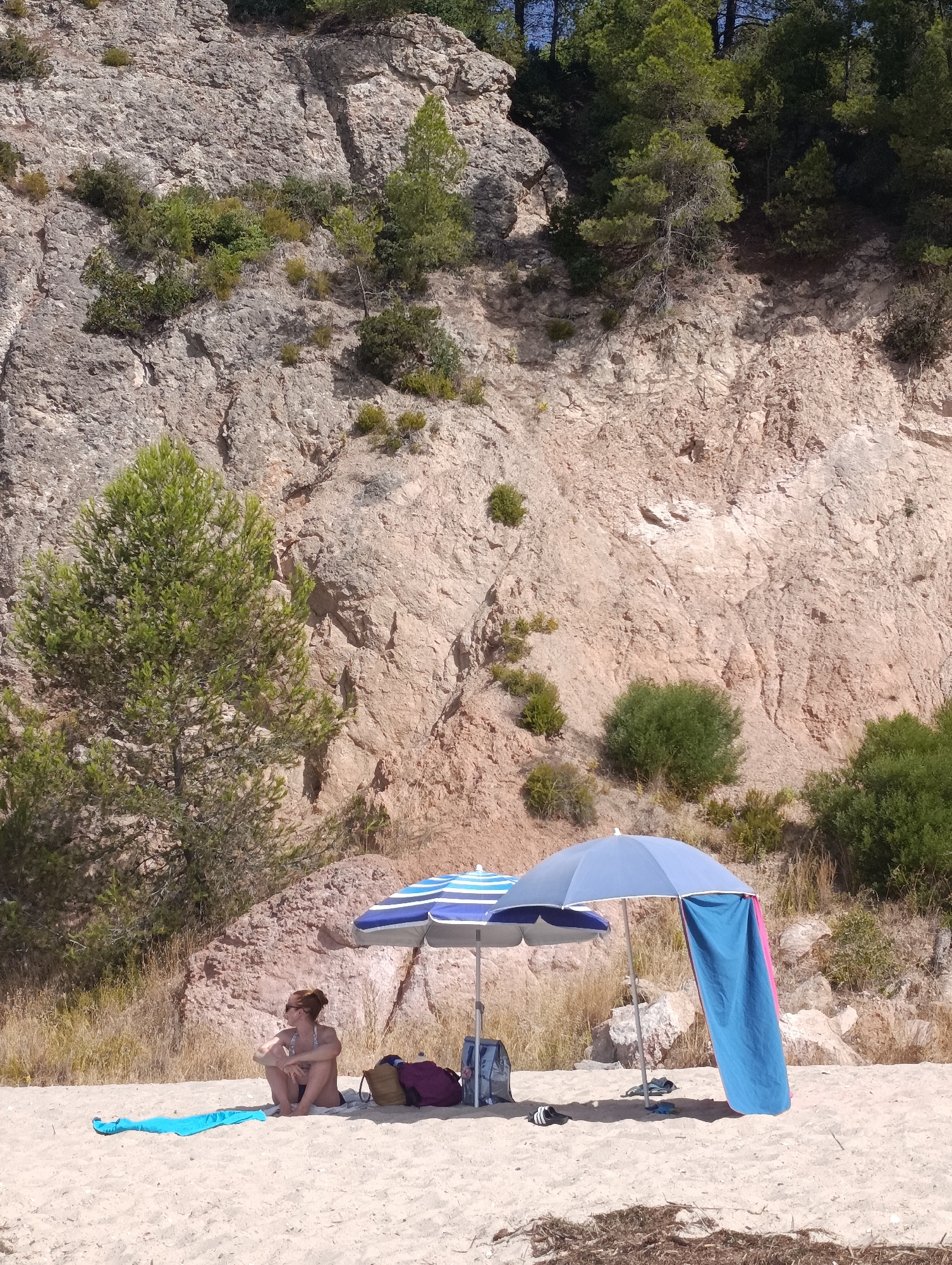 The image depicts a beach scene with a steep, rocky cliff face serving as a backdrop. In the foreground, a sandy beach is occupied by a person sitting in the shade of two umbrellas. The person, wearing a bikini, is looking to their left. Beside them are two beach bags, a blue towel, and a pair of flip-flops. A large blue umbrella has a towel draped over its side. The environment includes scattered vegetation on the cliff face and dry grass on the slope leading up to it. The scene suggests a sunny day at the beach. The location is not identifiable as being in the city of Setúbal, Portugal.