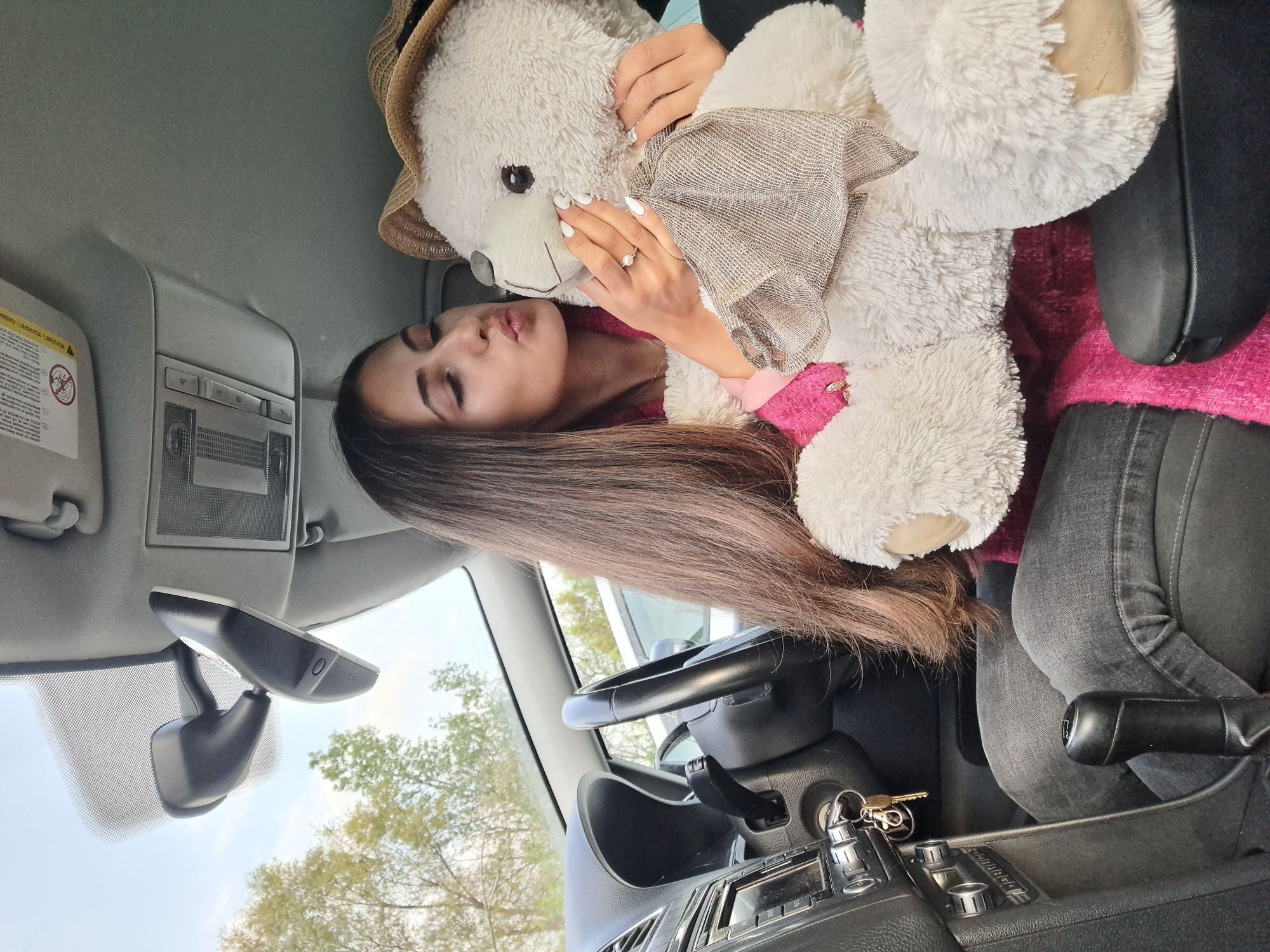 The image depicts a playful scene inside a car, presumably in Sofia, Bulgaria, during the day. A young woman with long, dark brown hair is the central figure, with her head tilted, eyes closed, and lips pursed as if blowing a kiss. She is holding a large, white, fluffy teddy bear close to her face.
The teddy bear is adorned with a straw hat and a patterned beige bow around its neck. The woman's hands gently cradle the bear, showcasing white nail polish and a ring with a spherical pearl-like detail on one finger. She is wearing a vibrant pink or magenta fluffy top and grey jeans.
The car's interior is visible, including a steering wheel, dashboard, and car keys in the ignition. An overhead console with buttons, a sun visor, and a rearview mirror are also prominent. Through the car windows, a bright sky and green trees indicate an outdoor, daytime setting with clear weather. On the sun visor, a safety warning label is visible; while the text is mirrored and mostly illegible, "WARNING" and various safety symbols can be discerned. The overall impression is one of affectionate interaction and personal comfort within a casual setting.